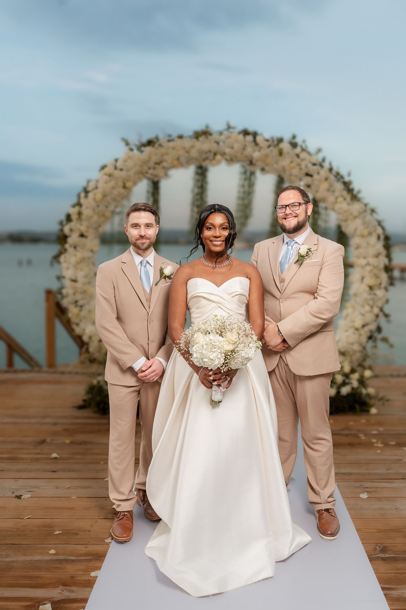 Bride in white gown holds bouquet flanked by two groomsmen in tan suits before circular floral arch near water.