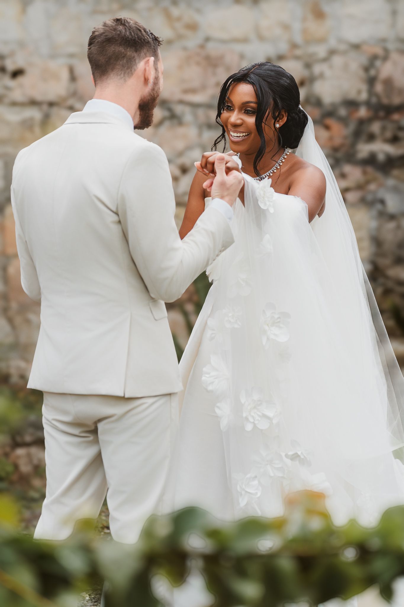 Interracial couple sharing a joyful moment during outdoor wedding ceremony, bride in floral gown, groom in cream suit.