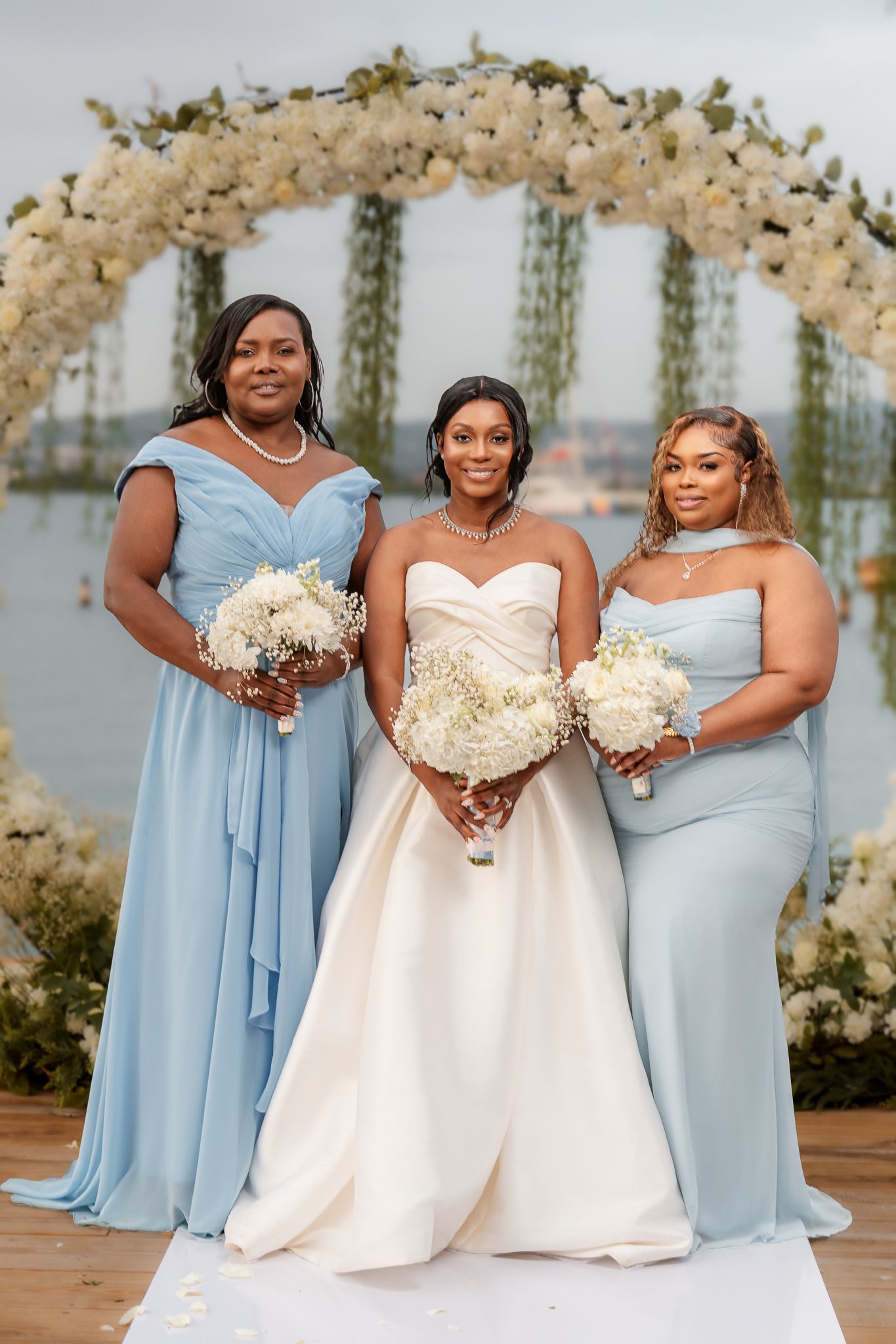 Bride in white gown poses with two bridesmaids in light blue dresses, holding white bouquets before a floral arch.