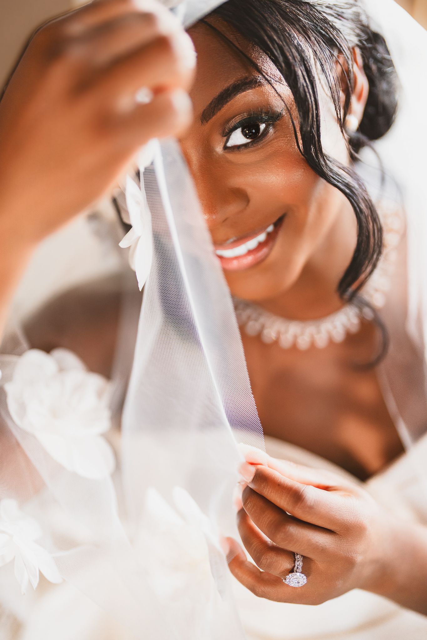 A radiant Black bride smiles while holding her lace veil, showcasing her diamond engagement ring and elegant pearl necklace.