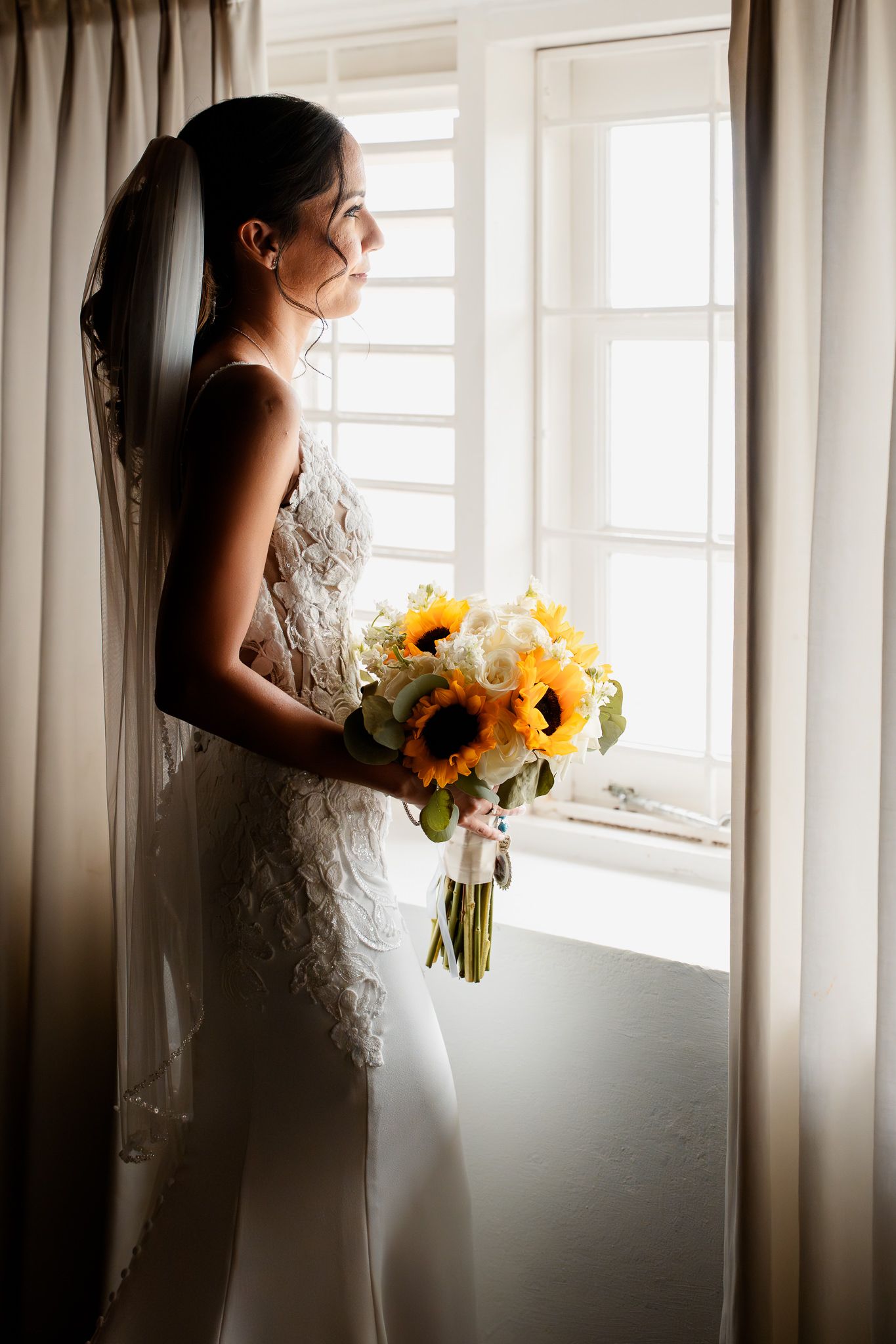 A bride in a white lace gown stands gracefully by a window holding a vibrant bouquet of sunflowers in natural light.