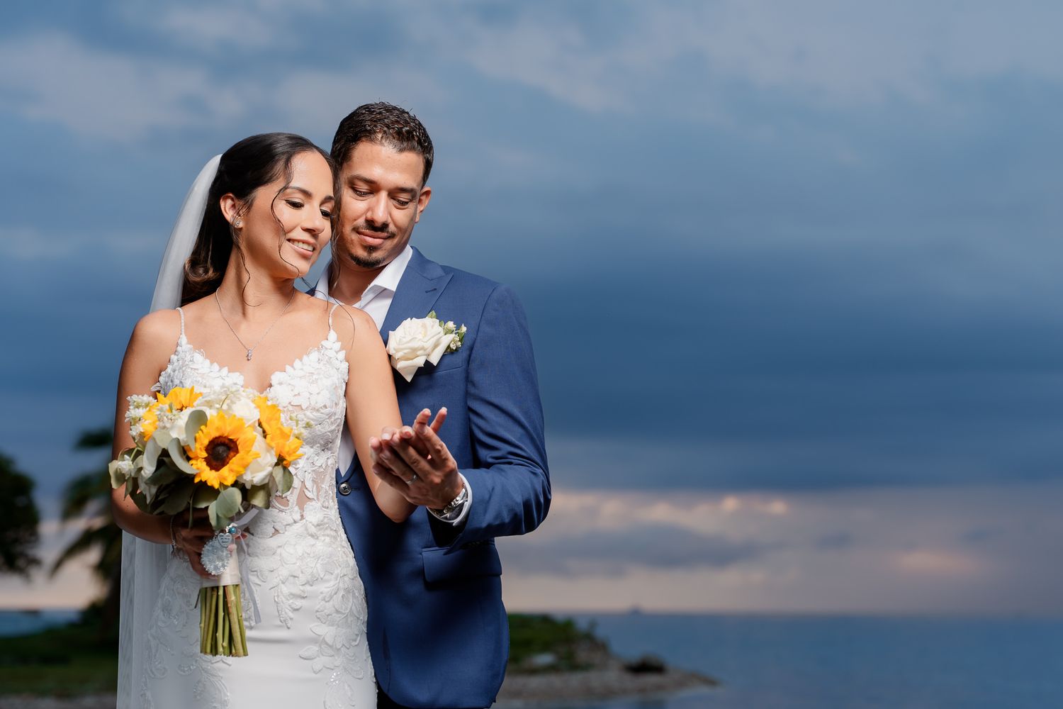 A couple shares a tender moment on their wedding day by the water, with sunflowers in the bridal bouquet.