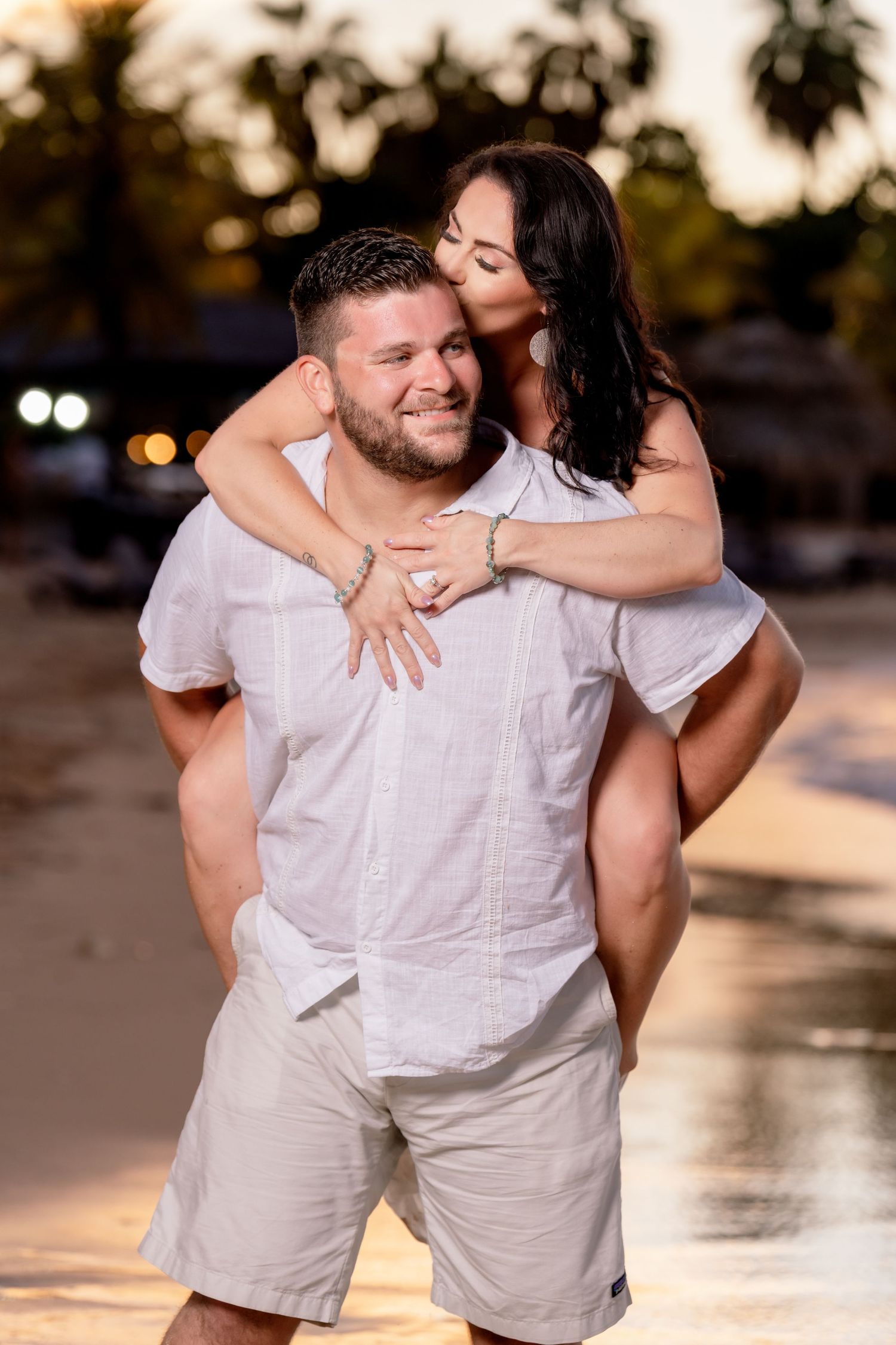 Happy couple embraces on tropical beach during golden hour sunset, with palm trees and glowing light in background.