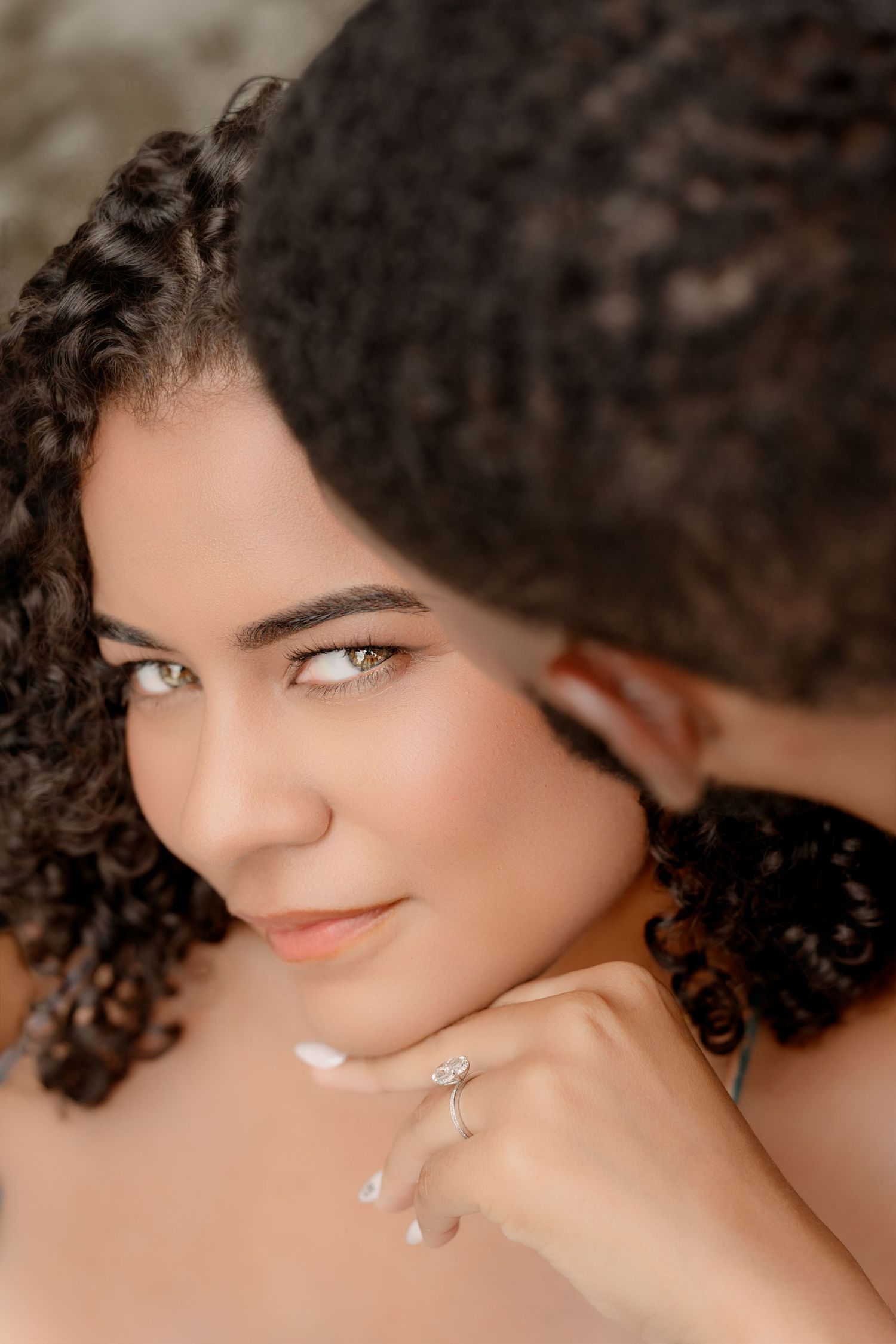 Close-up beauty portrait showing dark curly hair and a subtle smile against a soft beige background.