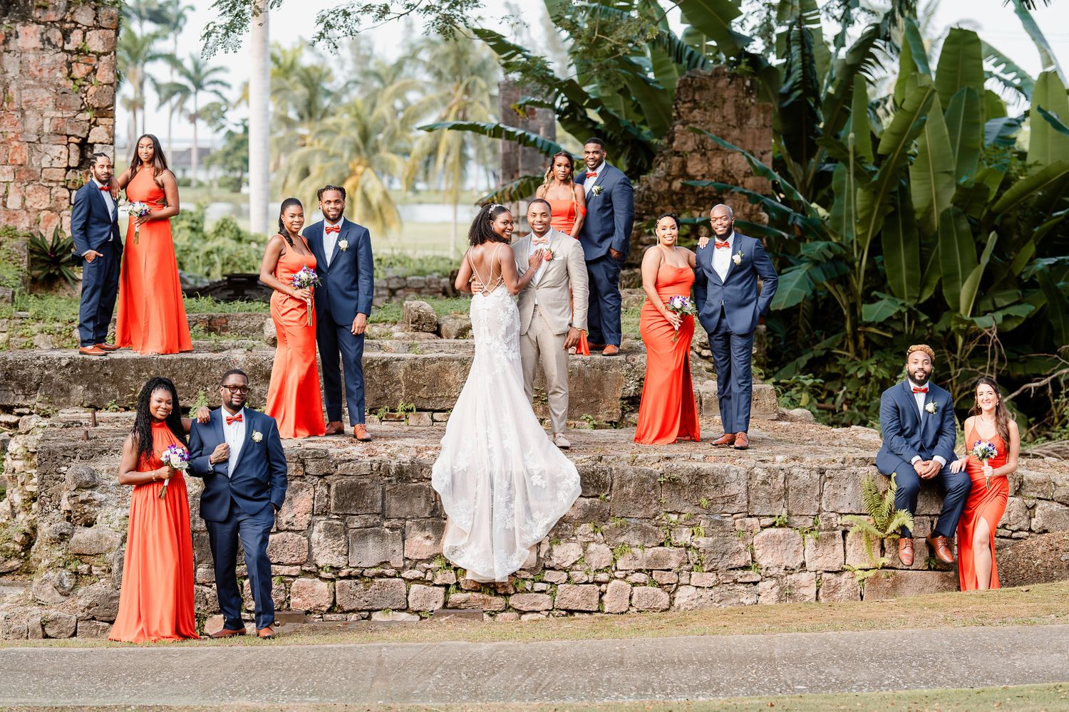 Wedding party in orange gowns and navy suits poses on stone ruins with tropical palms, bride and groom centered.