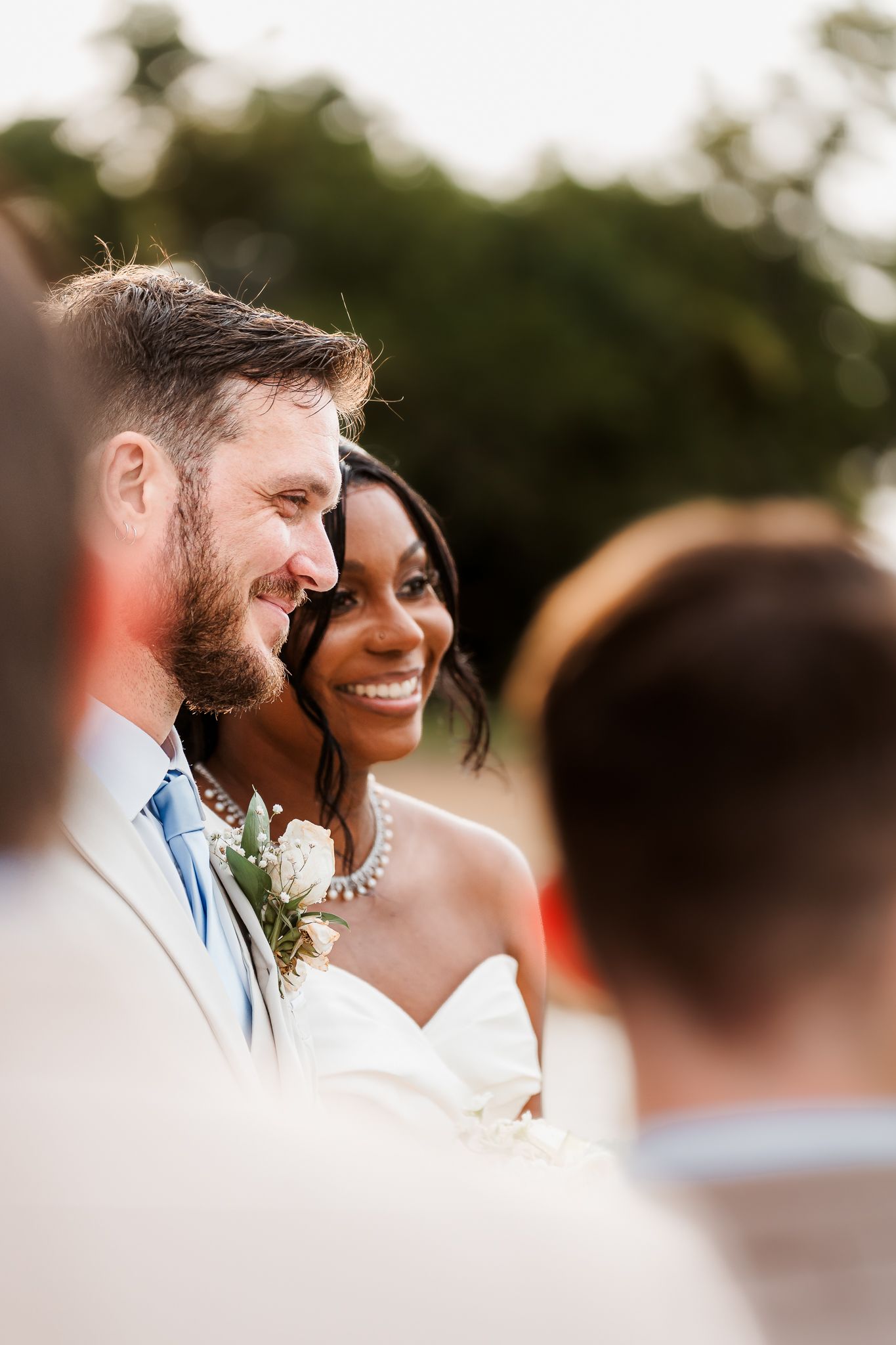 Interracial couple smiling joyfully during outdoor wedding ceremony, bride in white strapless gown with pearl necklace.