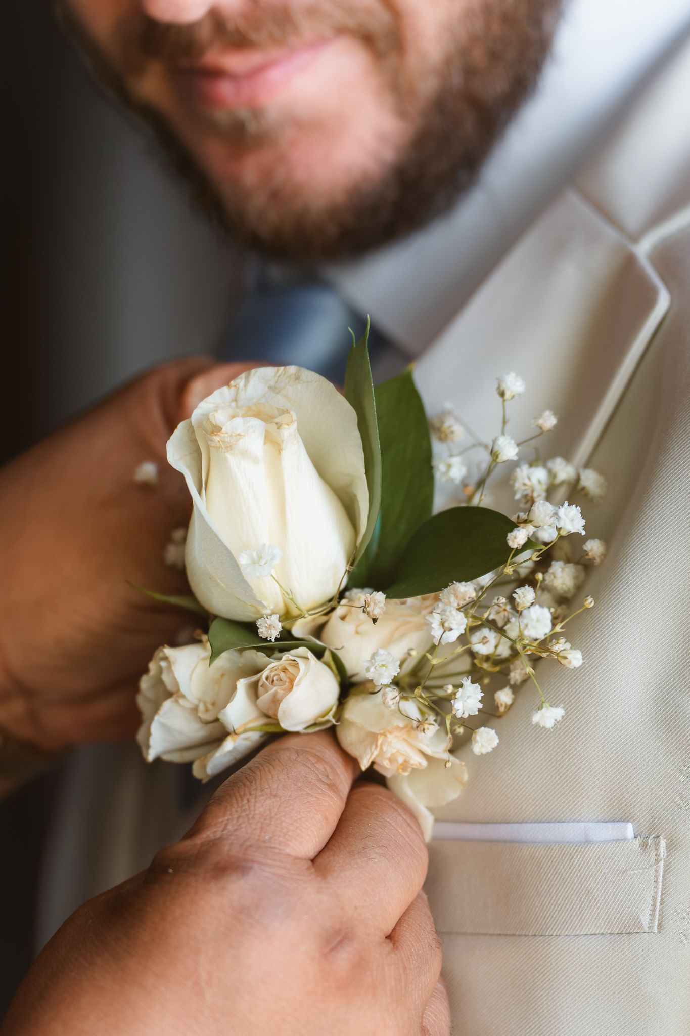 Close-up of a white rose boutonniere being pinned onto a beige suit lapel at a wedding ceremony.