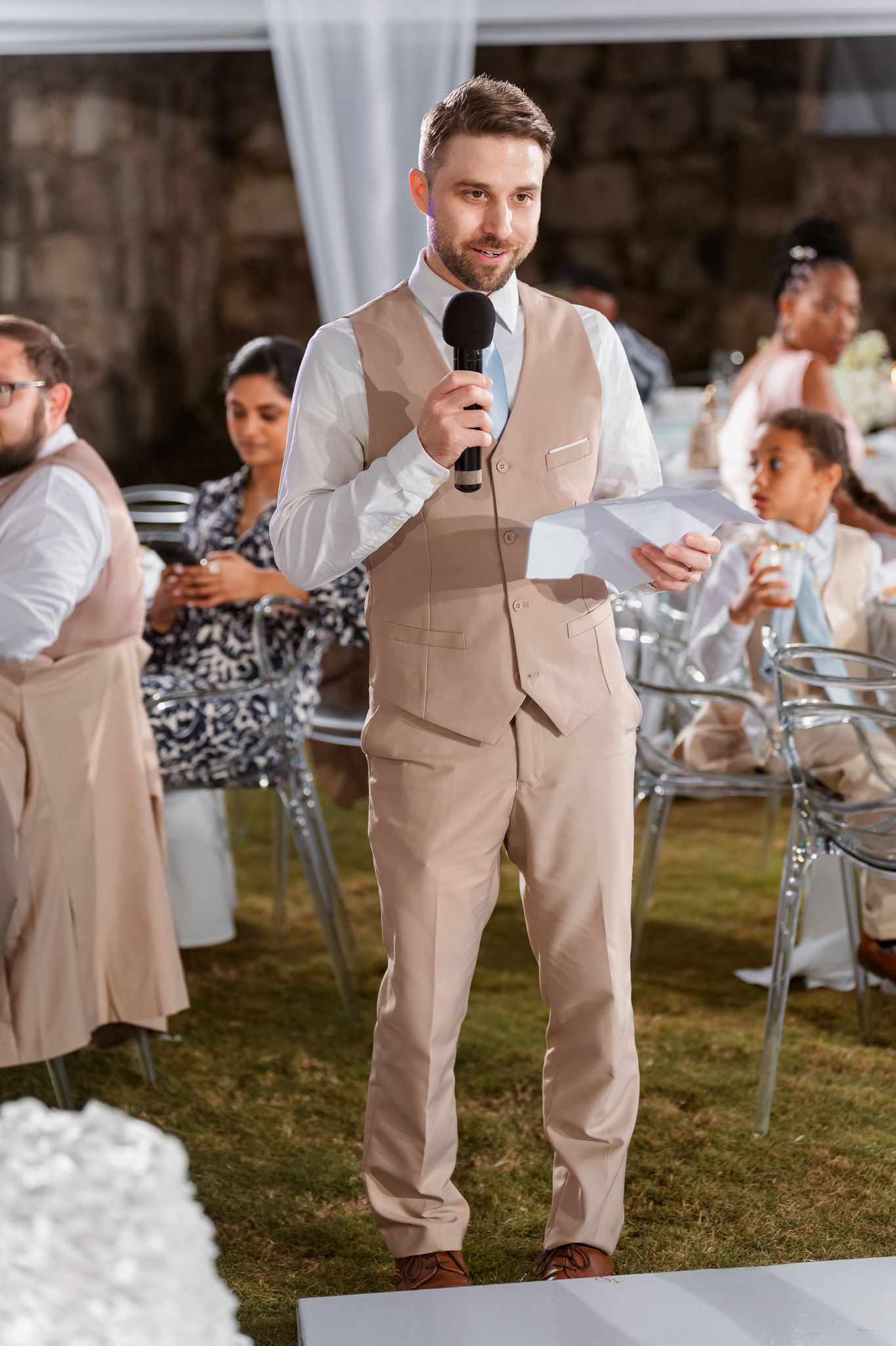A man in a beige suit delivers a speech at a wedding reception, holding a microphone and notes outdoors.