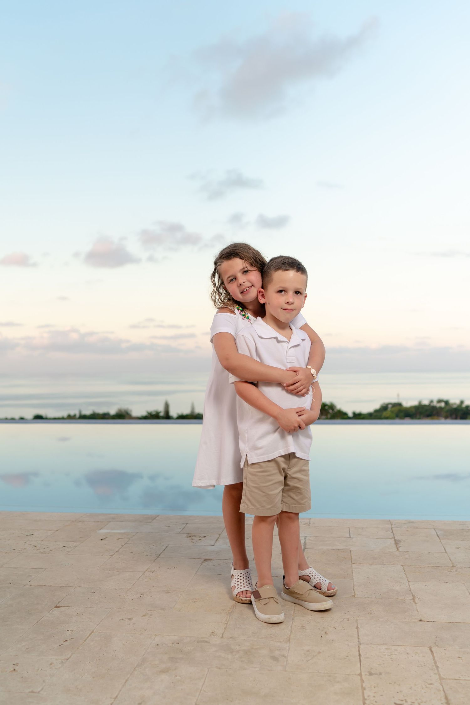 Two children embracing while standing on stone patio by infinity pool at sunset with ocean view in background.
