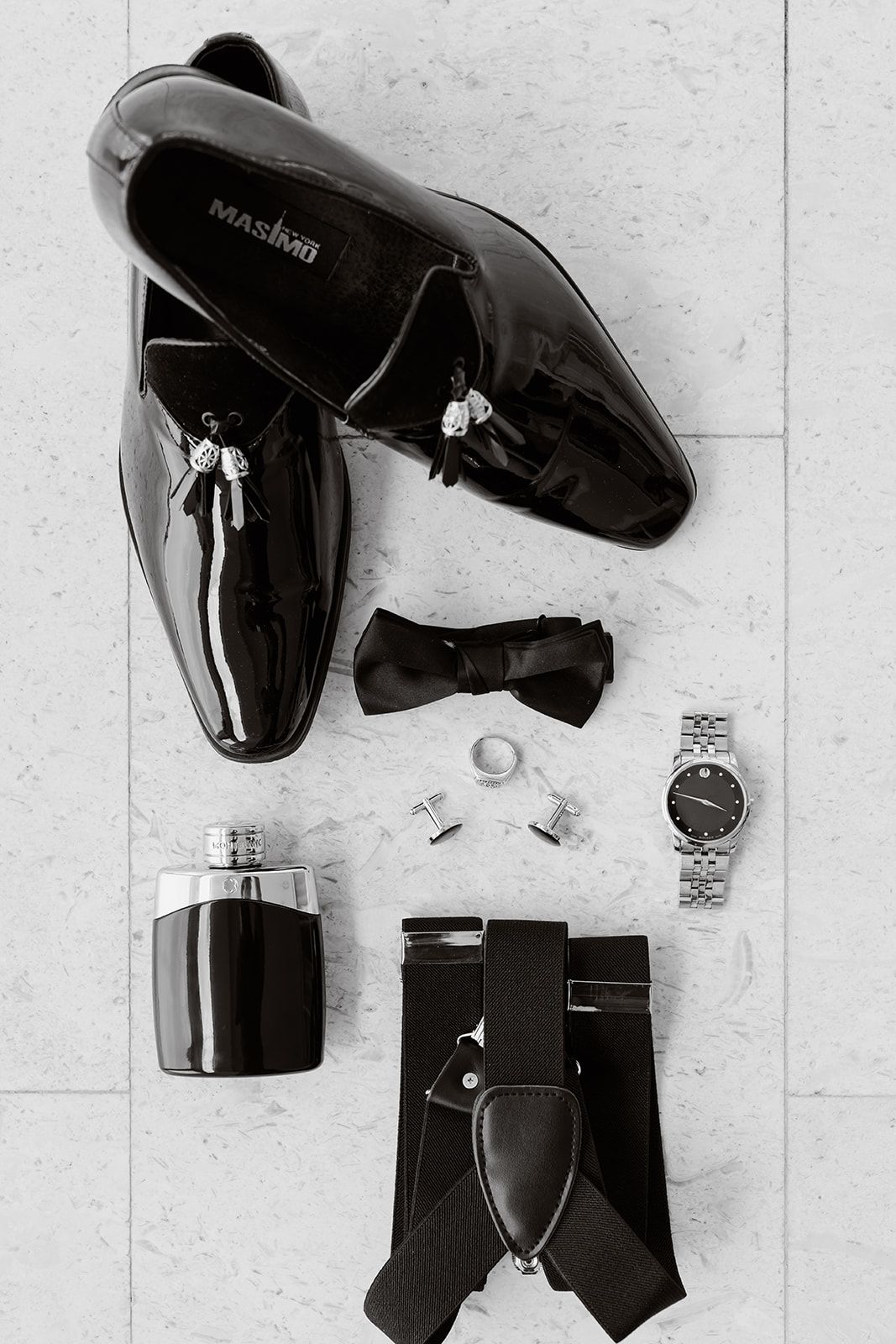 Black and white flatlay of formal men's accessories including patent leather shoes, bow tie, watch, cologne and suspenders.