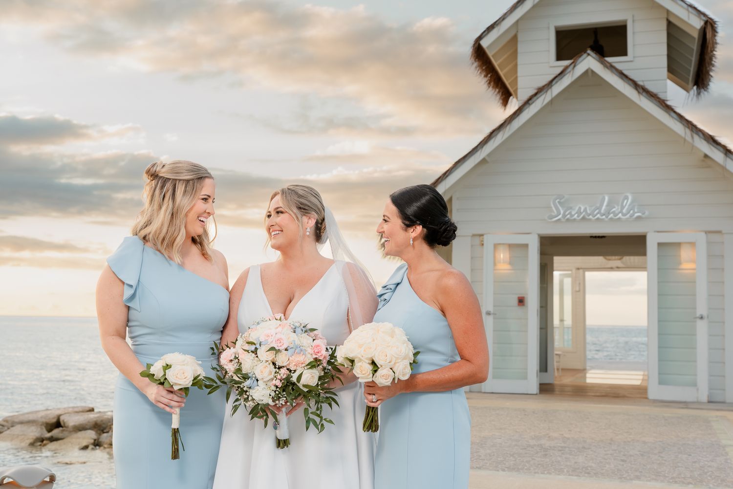 Bride and two bridesmaids in light blue dresses holding bouquets near a beachside chapel at sunset.