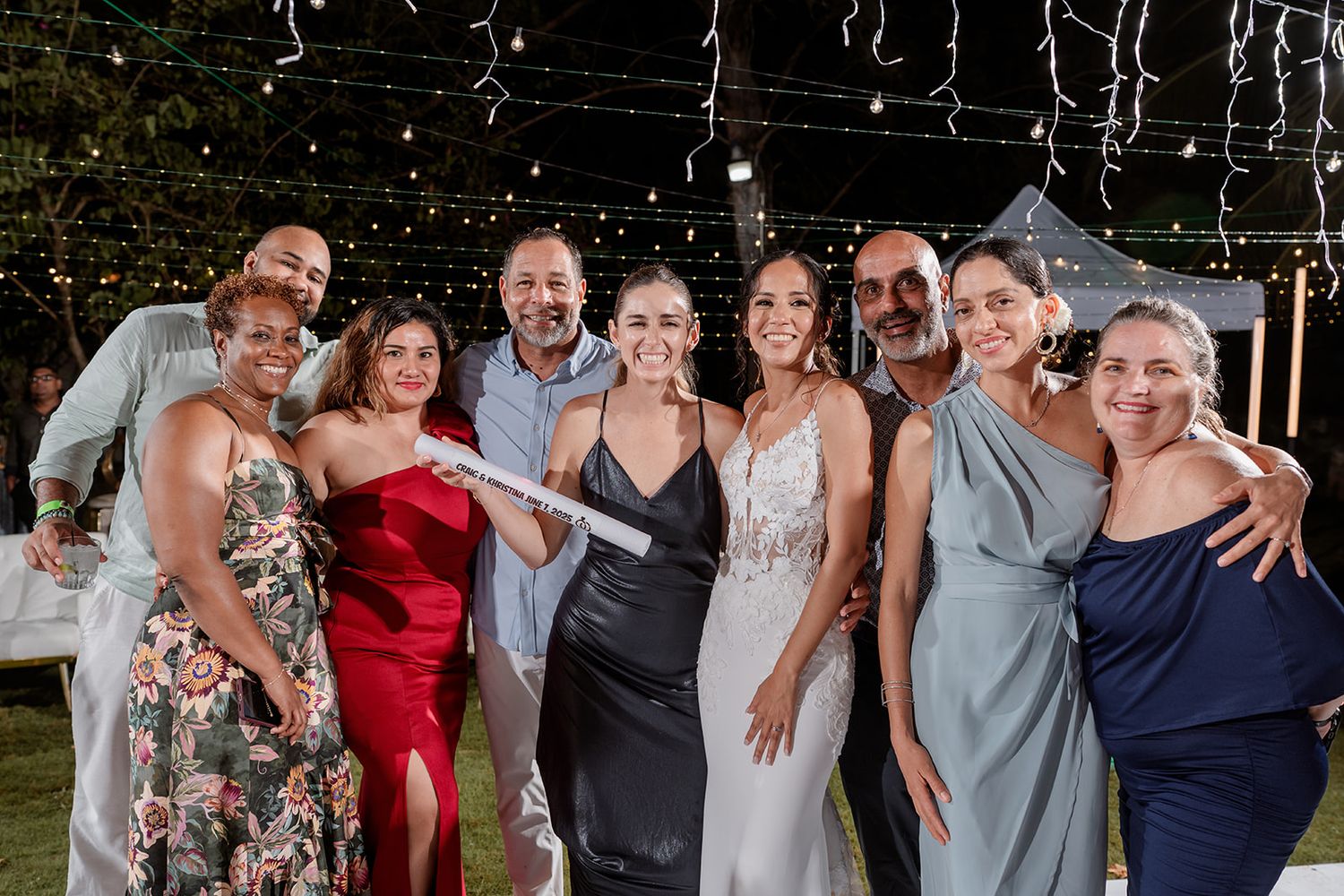 A group of wedding guests in formal attire smile together for photos at an outdoor evening reception.
