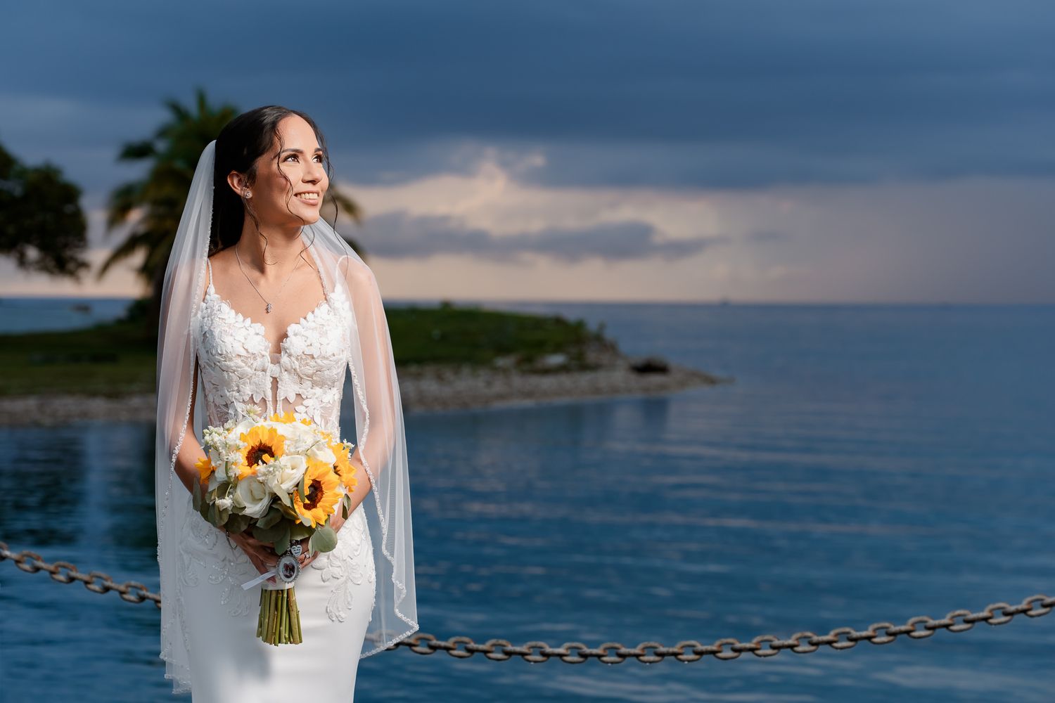 A bride in a white gown holds a sunflower bouquet while standing by the ocean at sunset with palm trees in background.