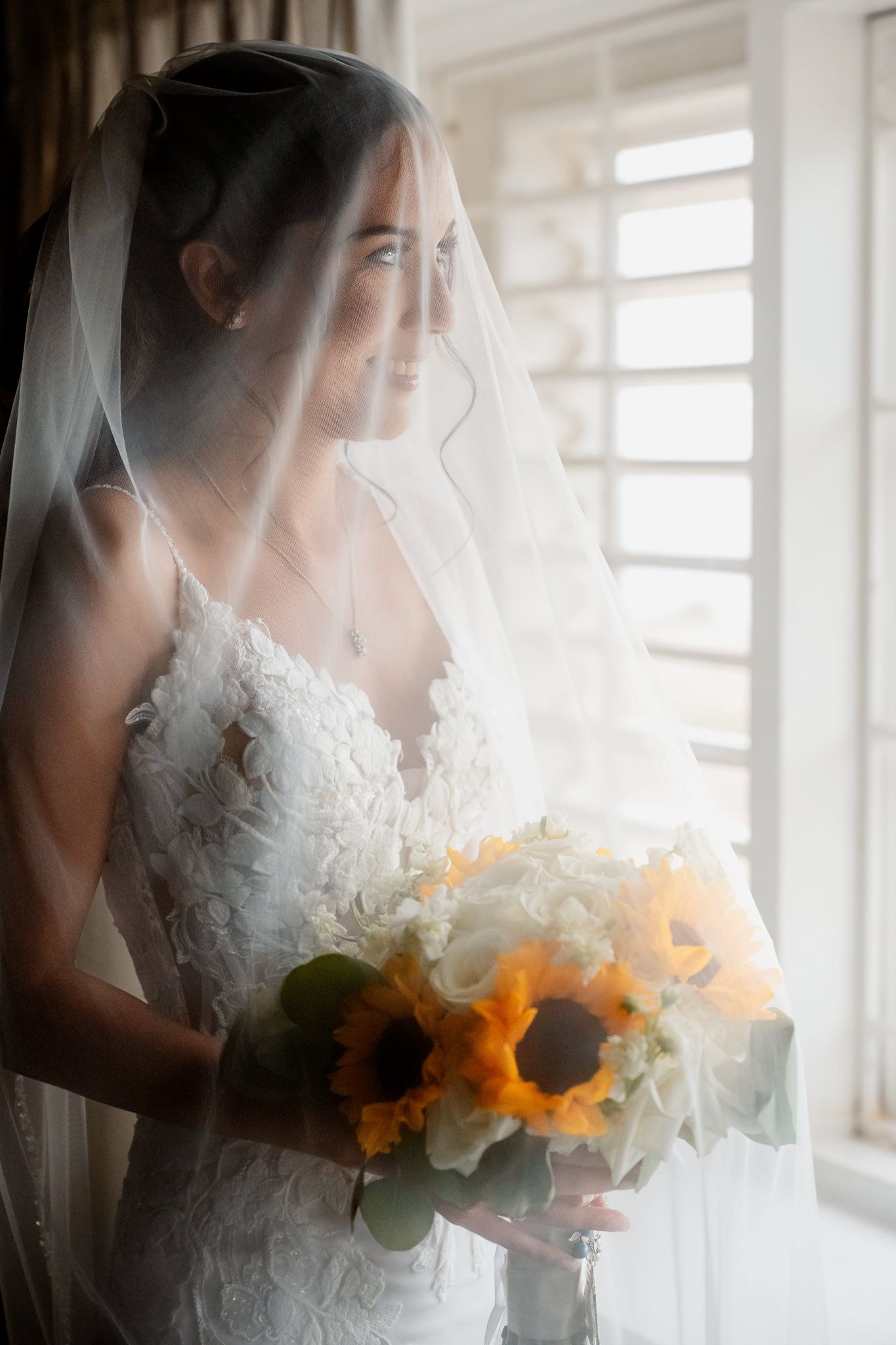 A bride in a white lace dress holds a sunflower bouquet while standing by a window with soft natural light.