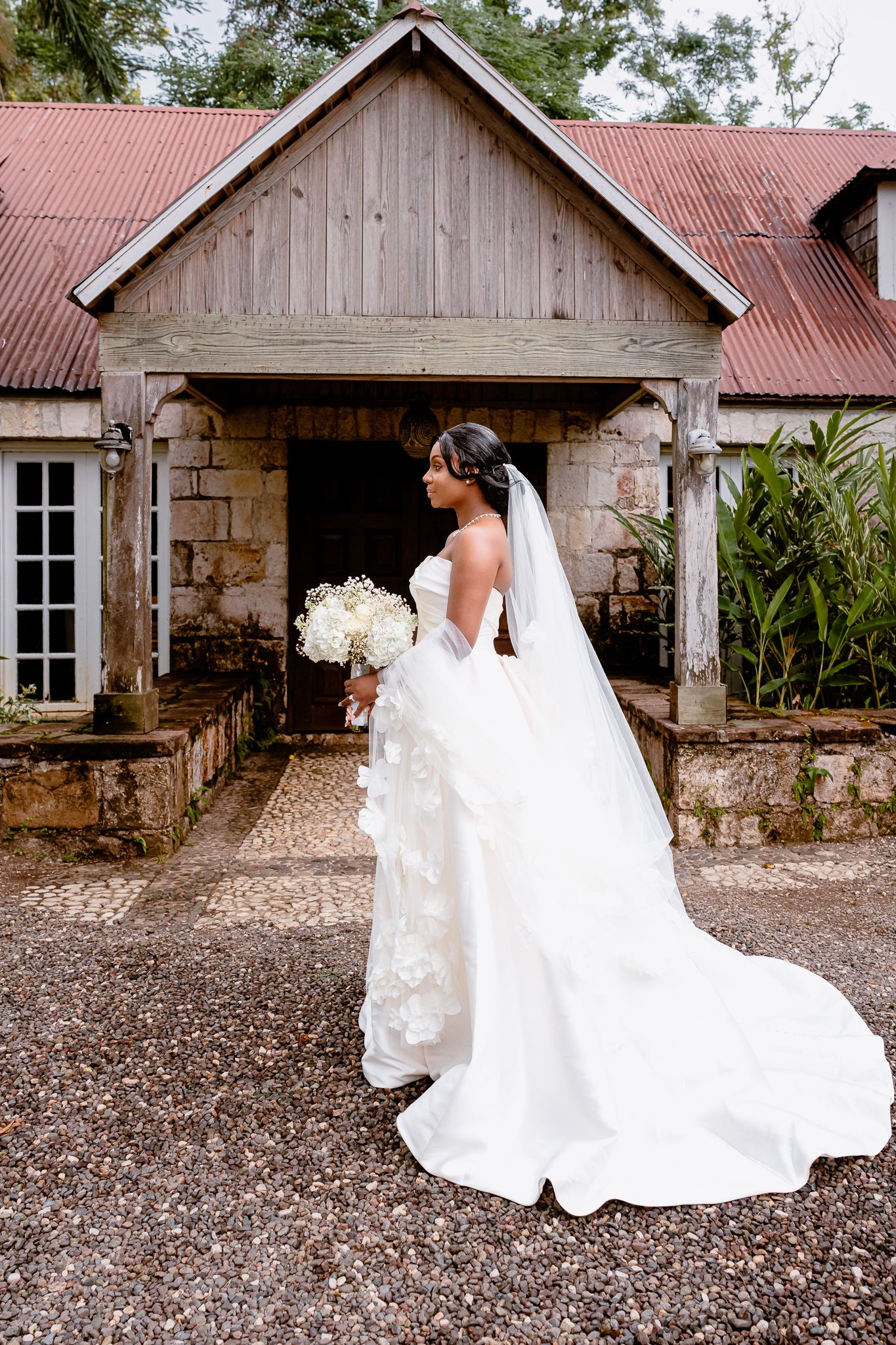 Bride in white gown and veil holds bouquet outside rustic stone building.