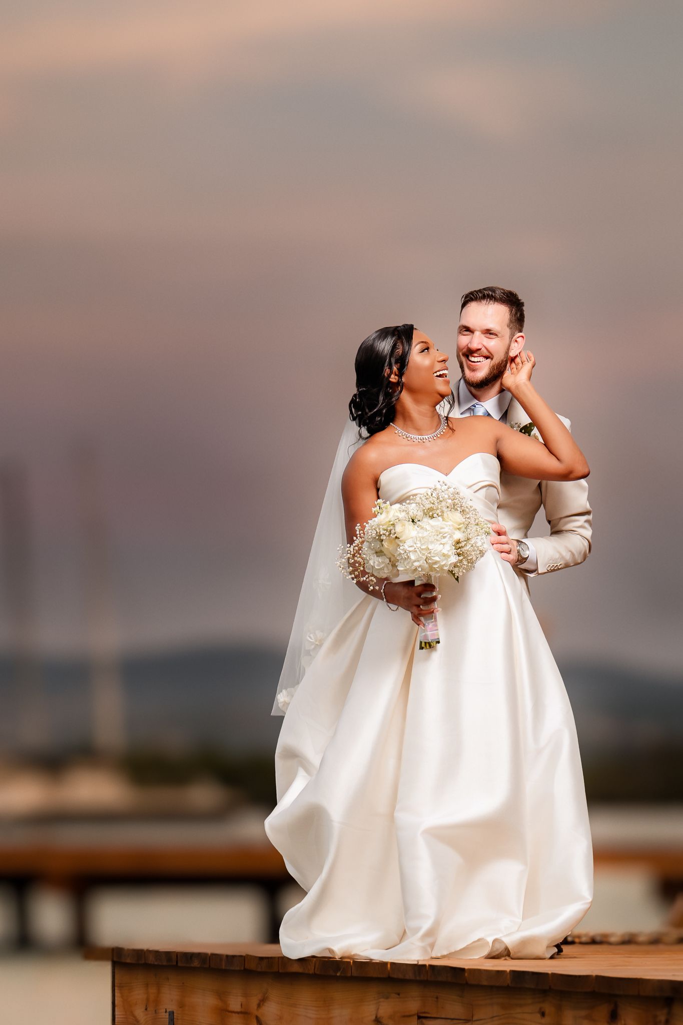 Interracial couple in wedding attire pose at sunset, bride in white gown holding bouquet, groom smiling joyfully.