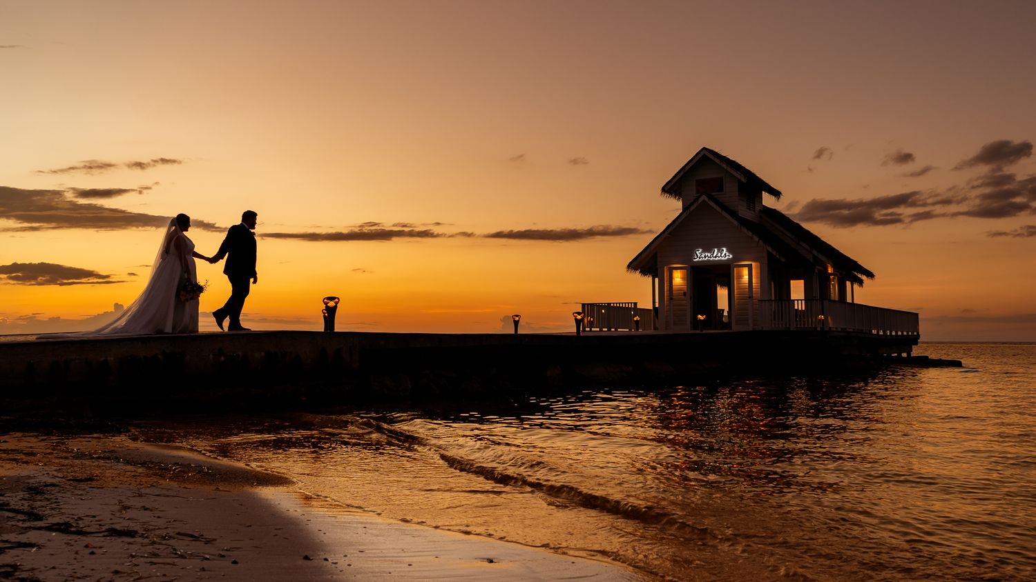 Silhouette of a wedding couple holding hands on a pier at sunset with a small overwater bungalow.