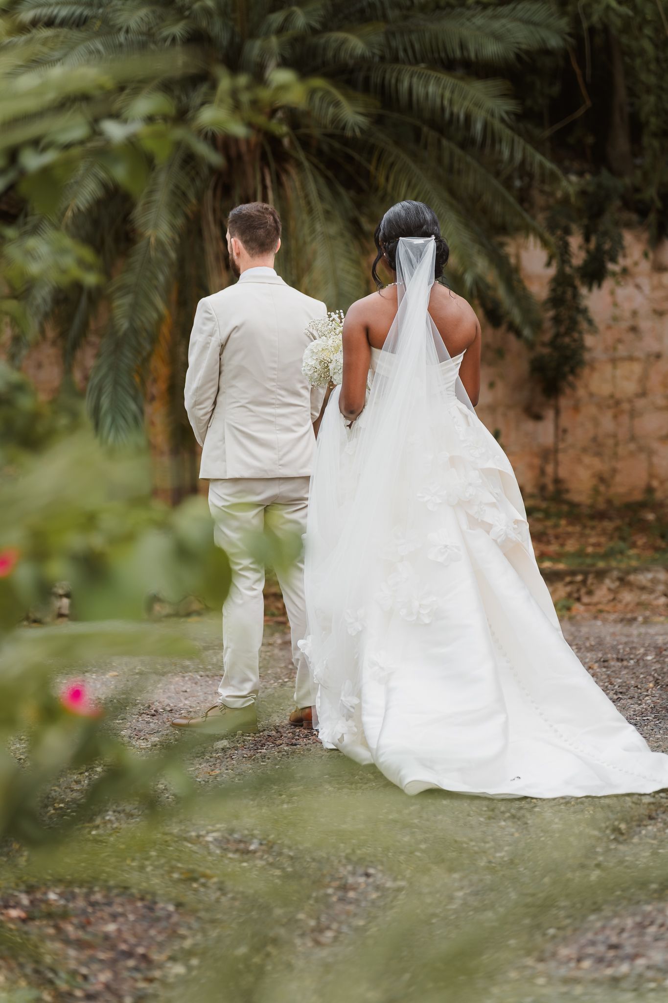 Bride in white gown with veil and groom in beige suit walk hand in hand through a lush garden path.