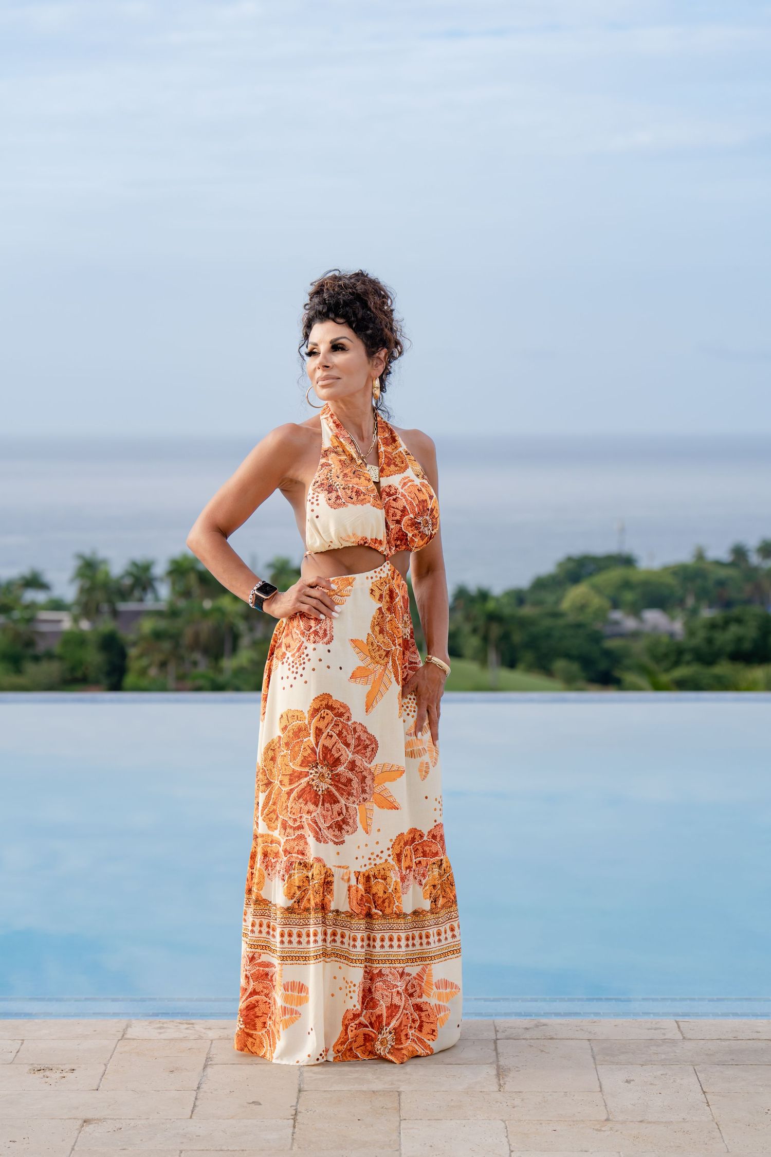 Person in a flowing orange and white floral maxi dress poses by an infinity pool overlooking a tropical coastal landscape.