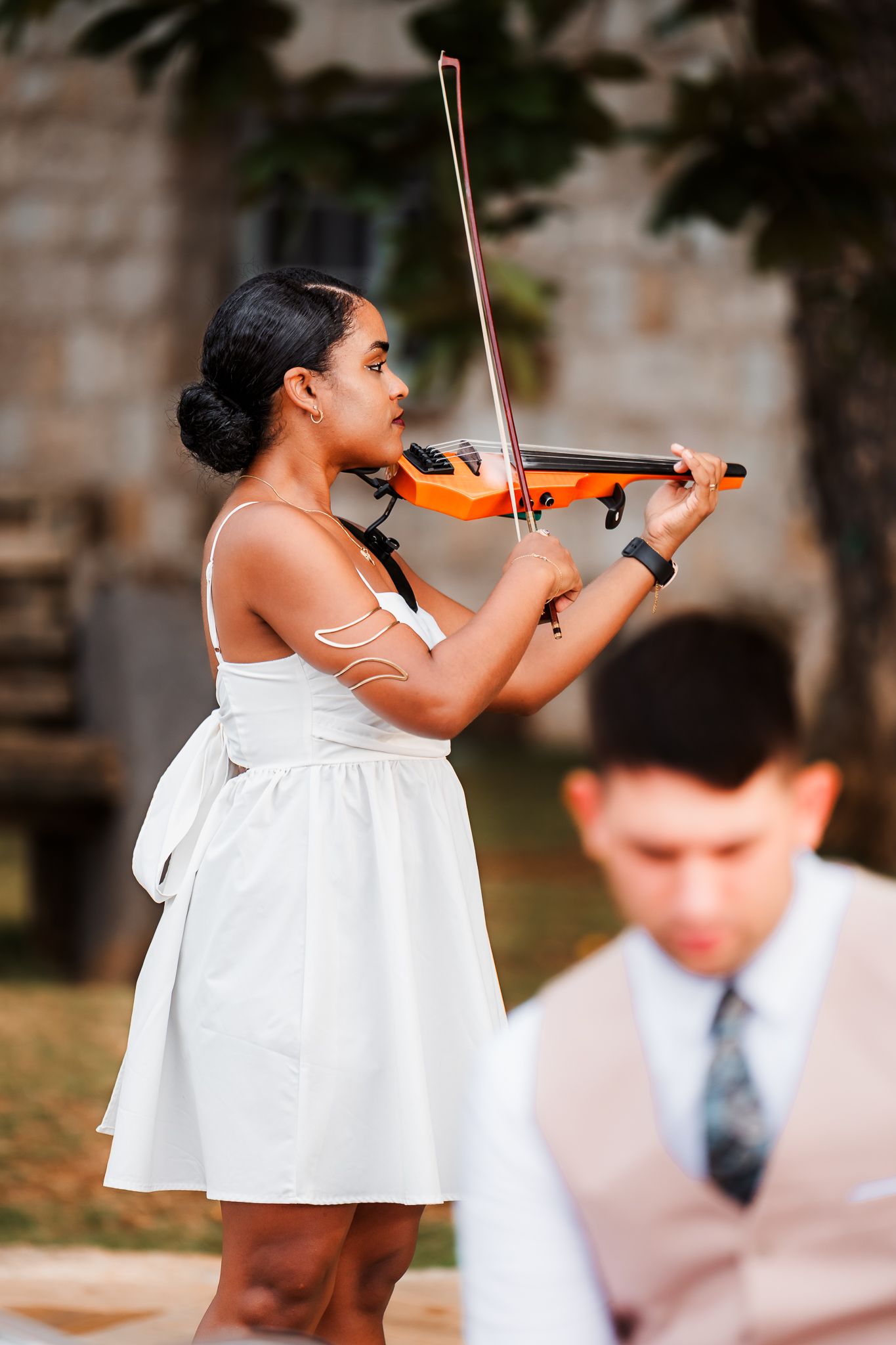 A woman in a white dress plays an orange violin outdoors, with a man in a suit visible in the foreground.