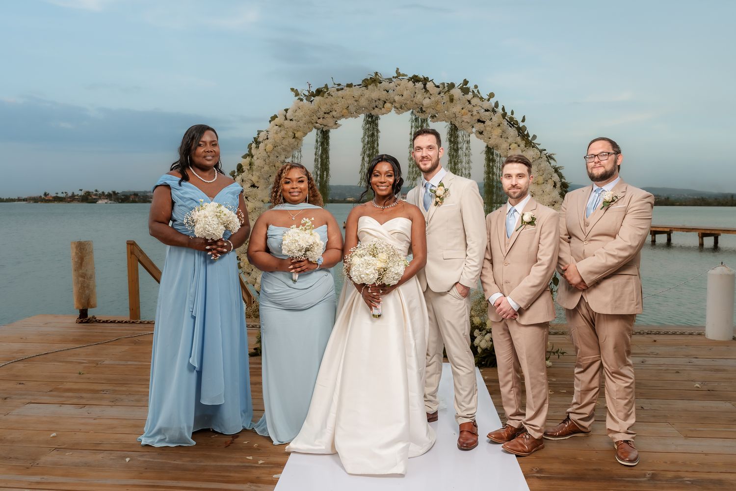 Wedding party poses on waterfront dock with floral arch, bride in white gown, bridesmaids in blue, groomsmen in tan suits.