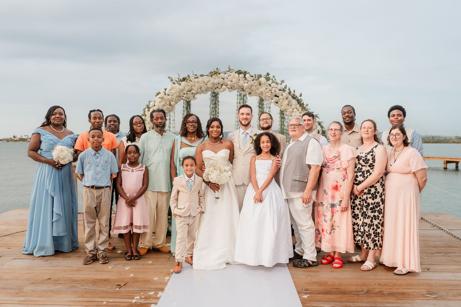 Wedding party poses on waterfront dock with floral arch, bride and groom centered among family in formal attire.