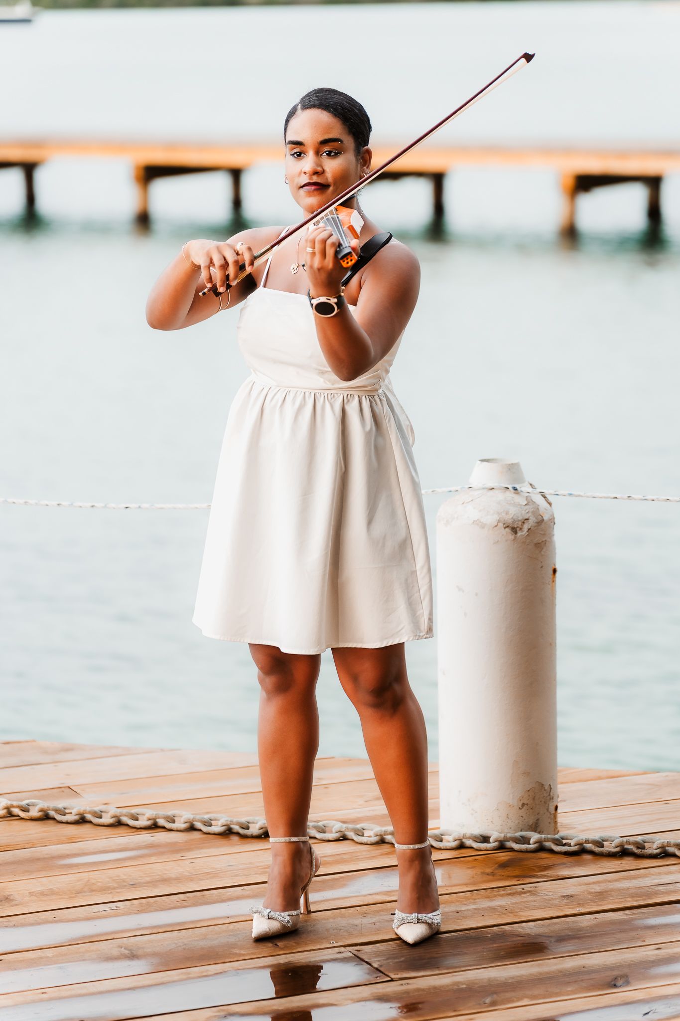 Woman in white dress playing violin on a wooden dock by the water.