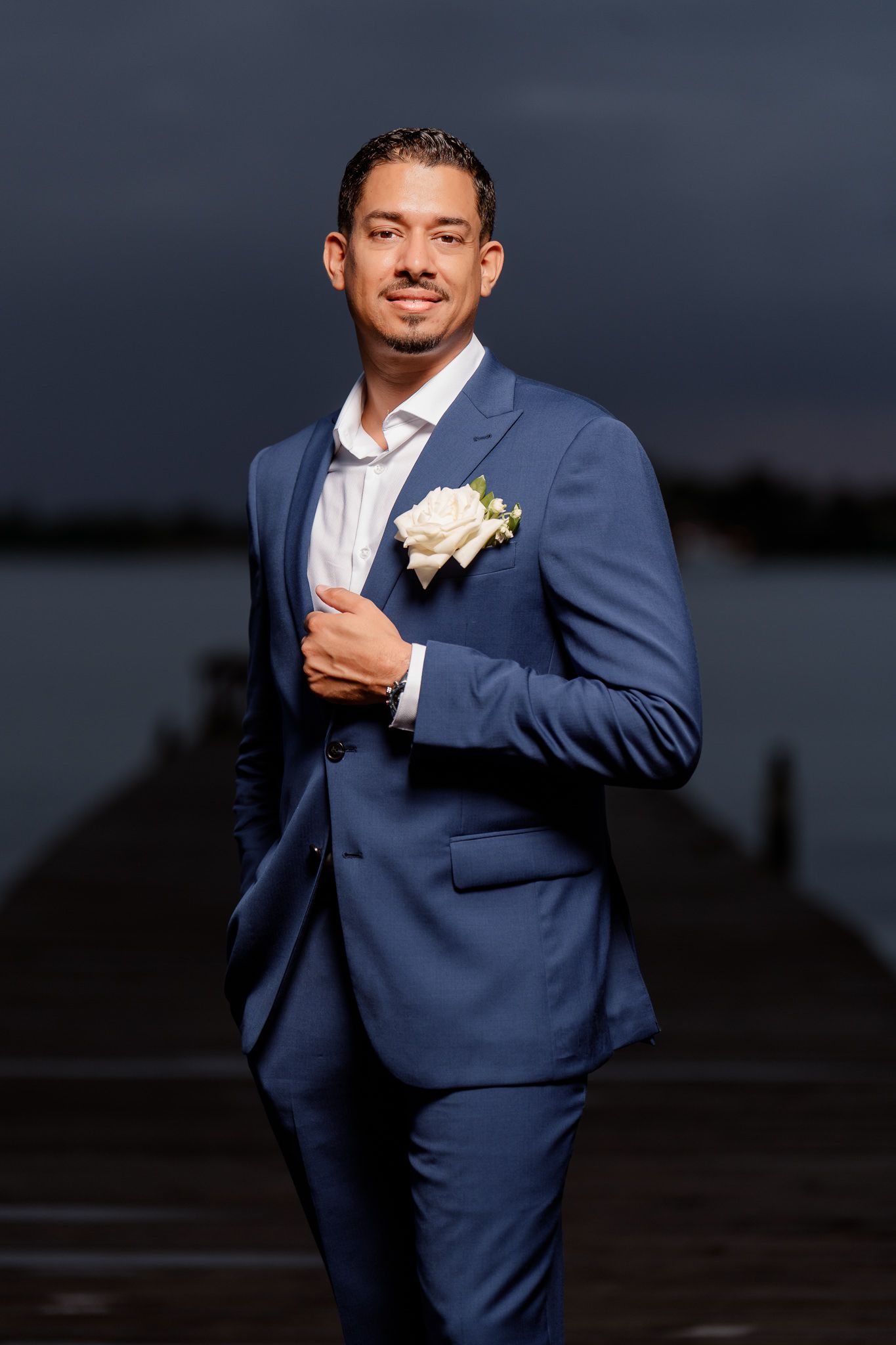 A groom in a navy blue suit and white boutonniere poses outdoors at dusk for a formal wedding portrait.