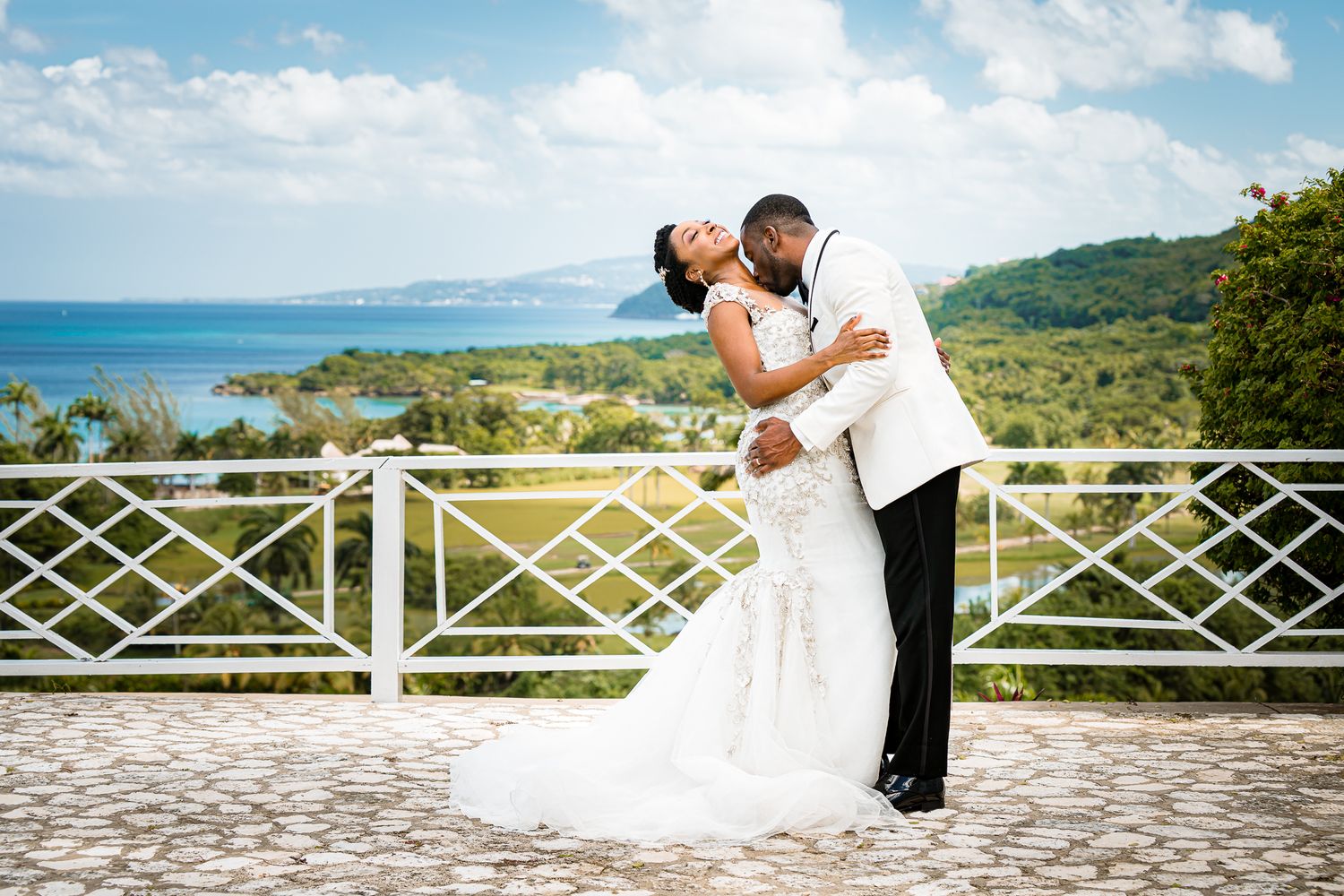 A bride and groom share a romantic kiss at an outdoor wedding venue overlooking a tropical ocean landscape.