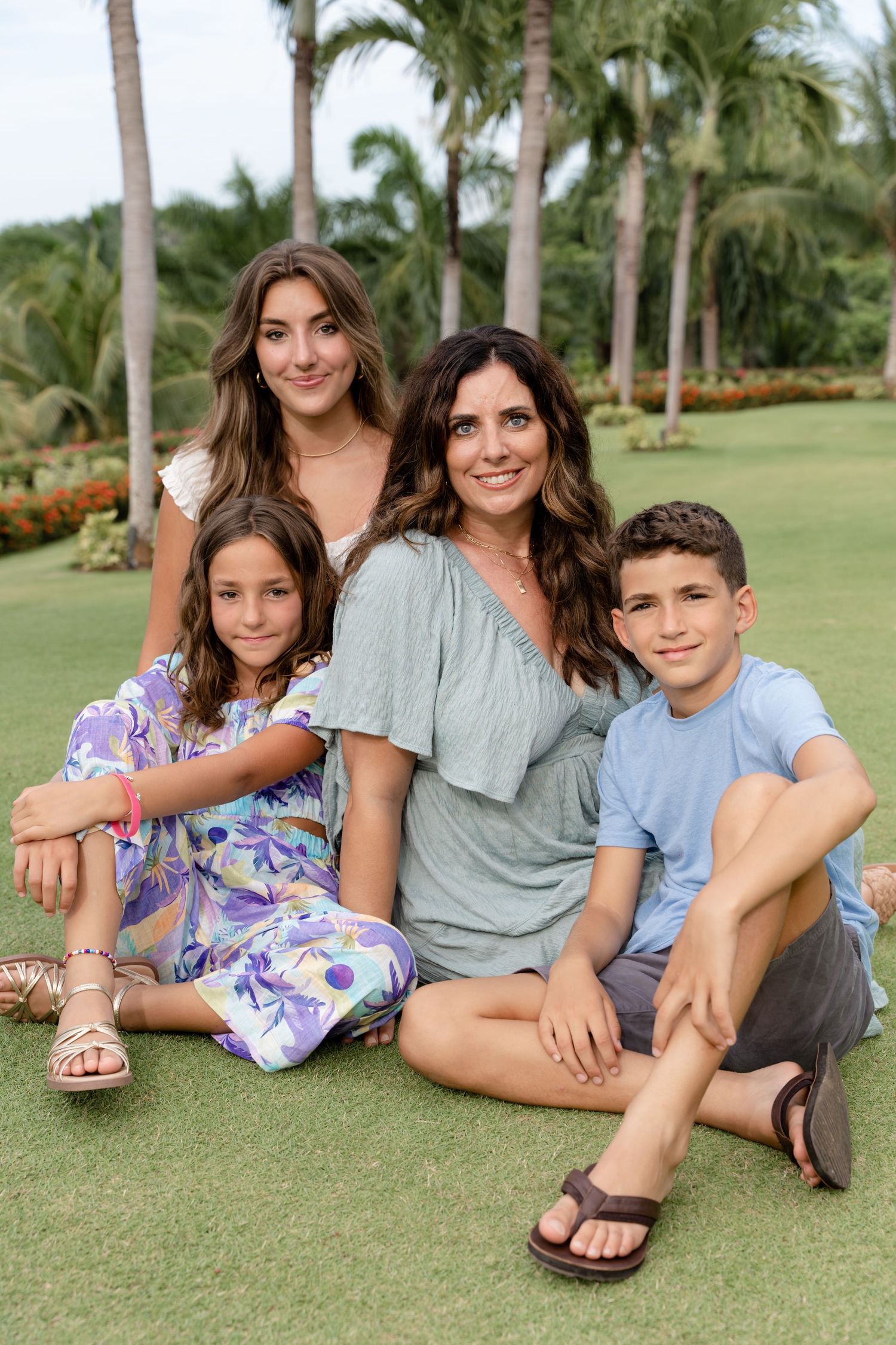 A group sits casually together on green grass with tall palm trees in the background of a tropical setting.