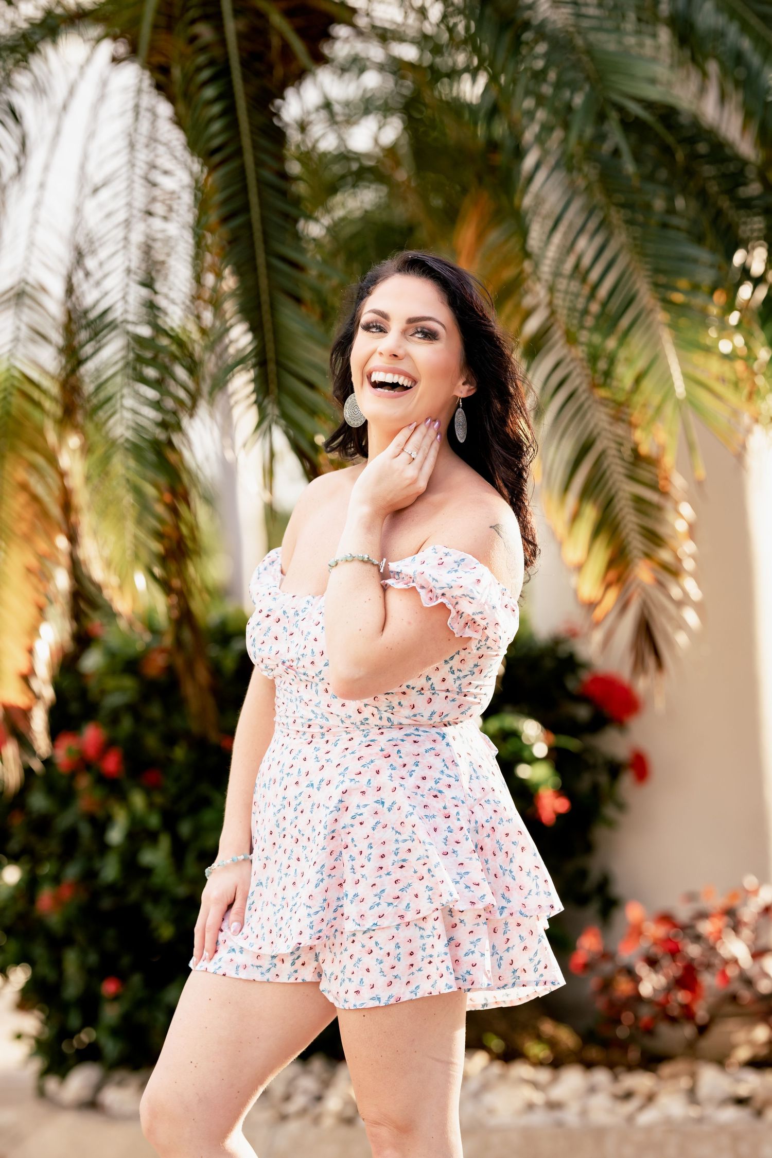 Woman in floral print off-shoulder romper laughs joyfully while posing against tropical palm fronds in outdoor setting.