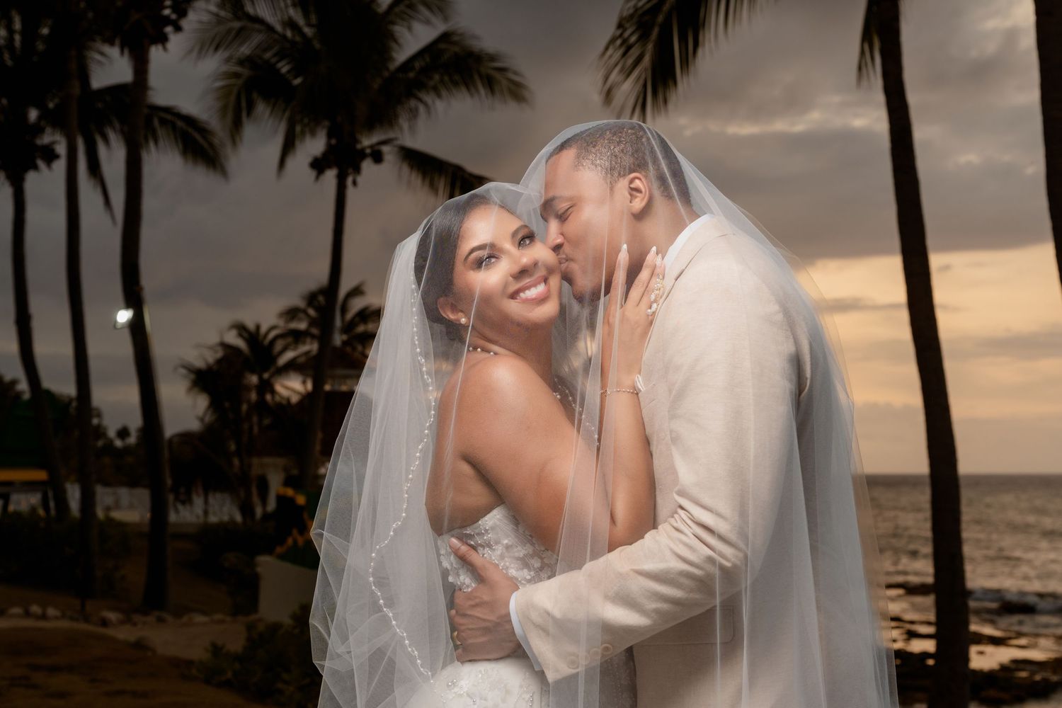A newlywed couple shares an intimate moment at sunset on a tropical beach with palm trees lining the shore.