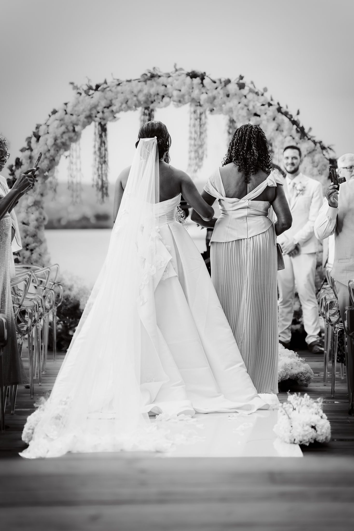 Black and white photo of a bride walking down the aisle with bridesmaid toward a floral arch.