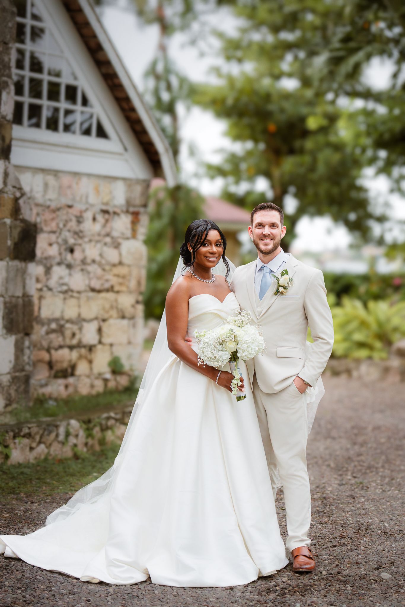 Interracial couple poses in wedding attire near stone building, bride in white gown holding bouquet, groom in beige suit.