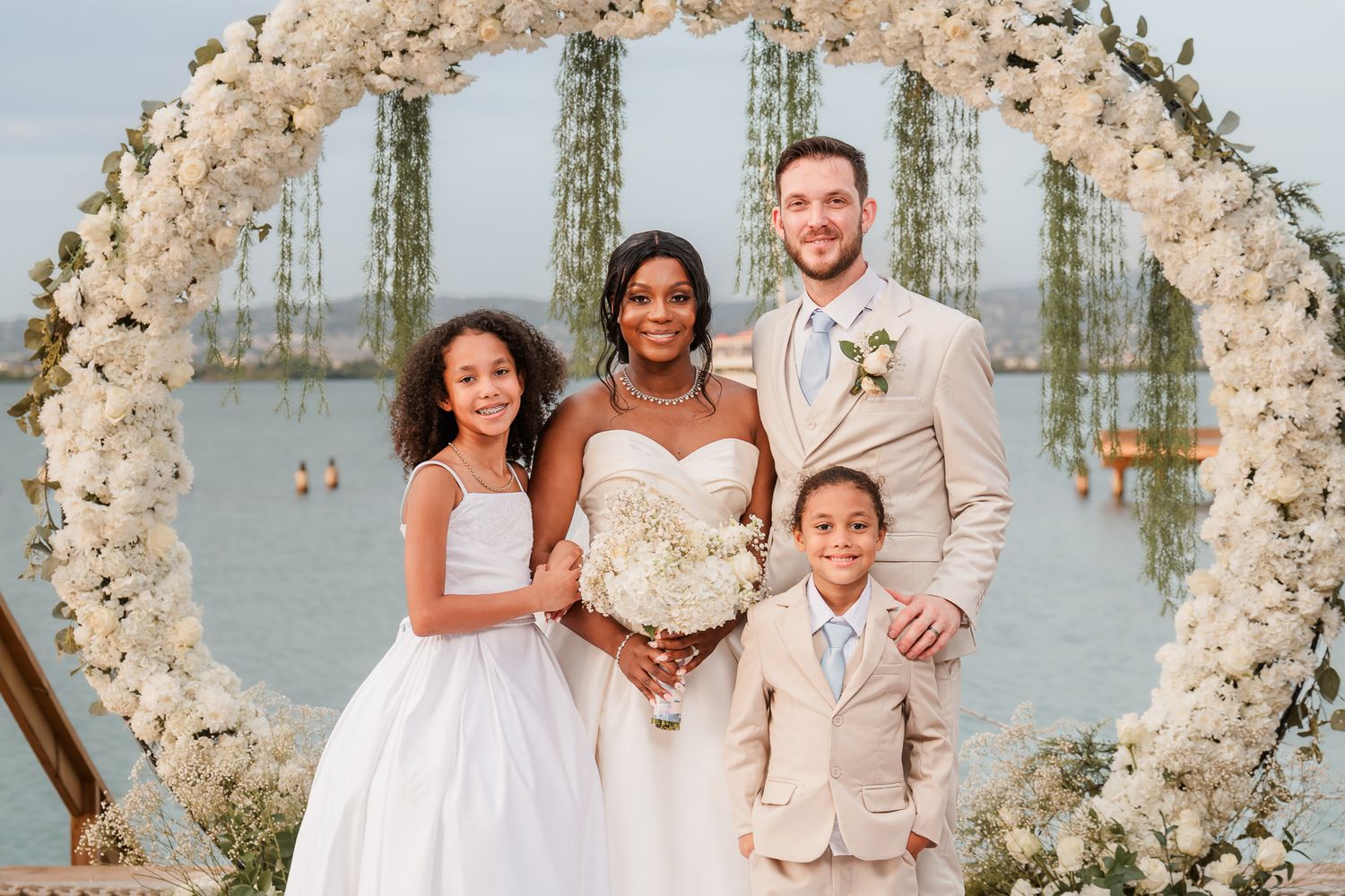 A bride and groom pose with two children in front of a floral arch at an outdoor waterfront wedding ceremony.