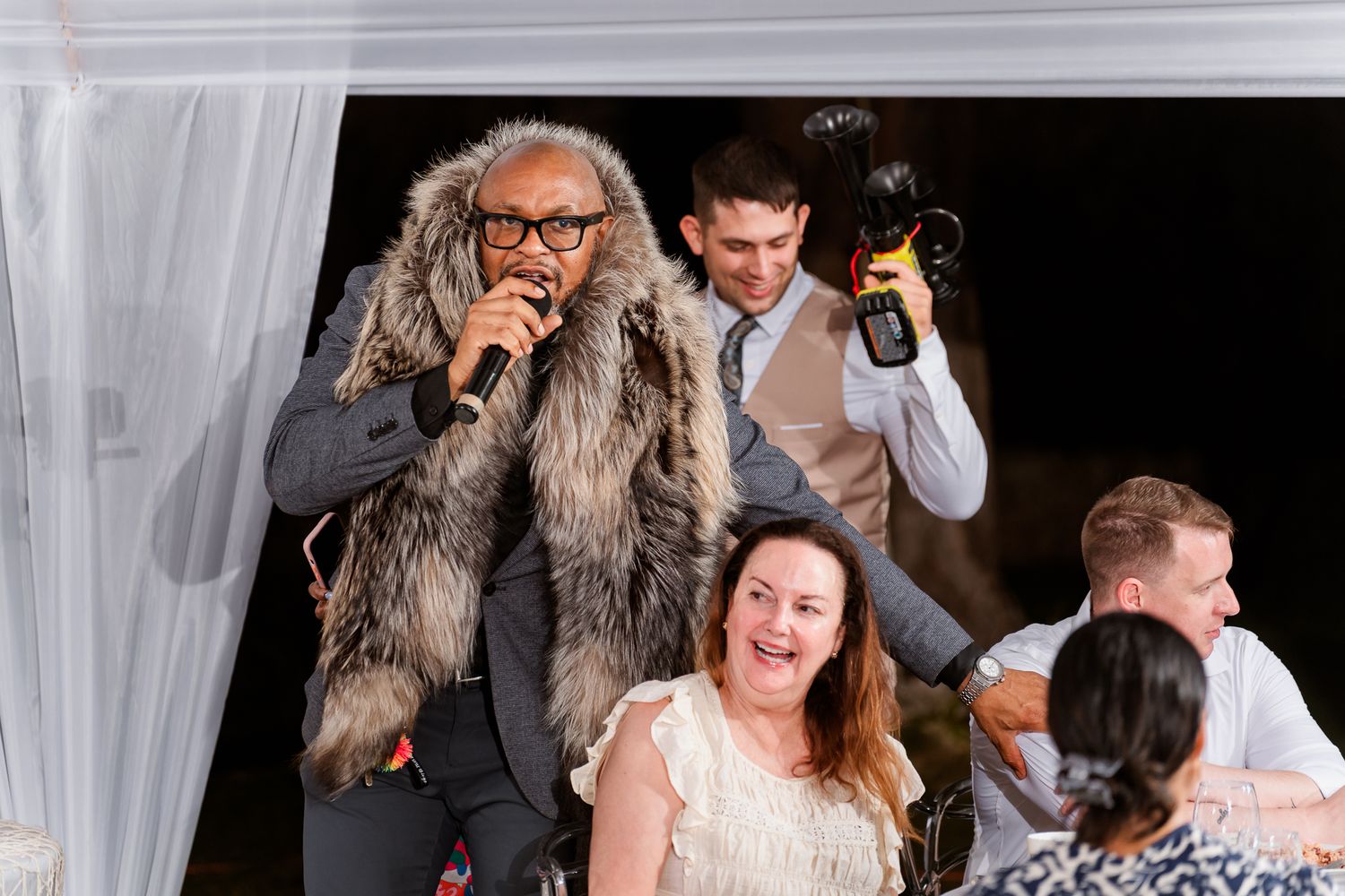 Performer in fur vest sings into microphone at wedding reception while guests laugh and photographer captures moments.
