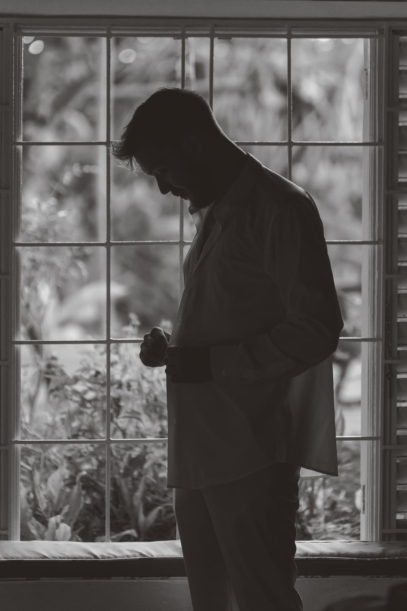 Black and white silhouette of a man buttoning his shirt in front of a large window with garden views.