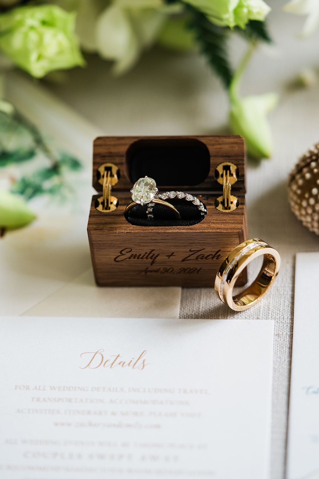 Wedding rings displayed in a personalized wooden box with Emily and Zach engraving next to floral decor and details card.