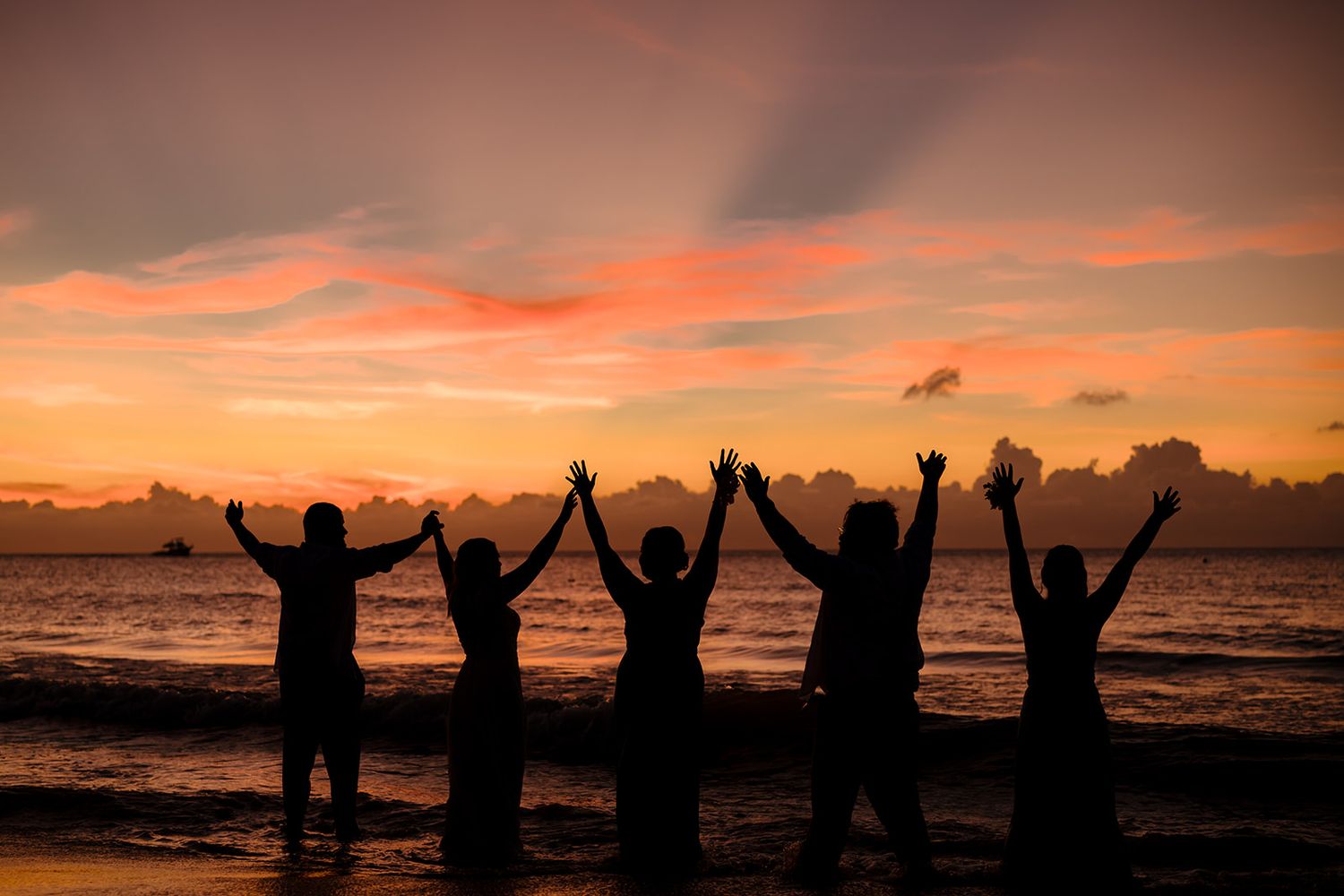 Silhouettes of people celebrating with raised arms against a vibrant orange sunset at the beach.