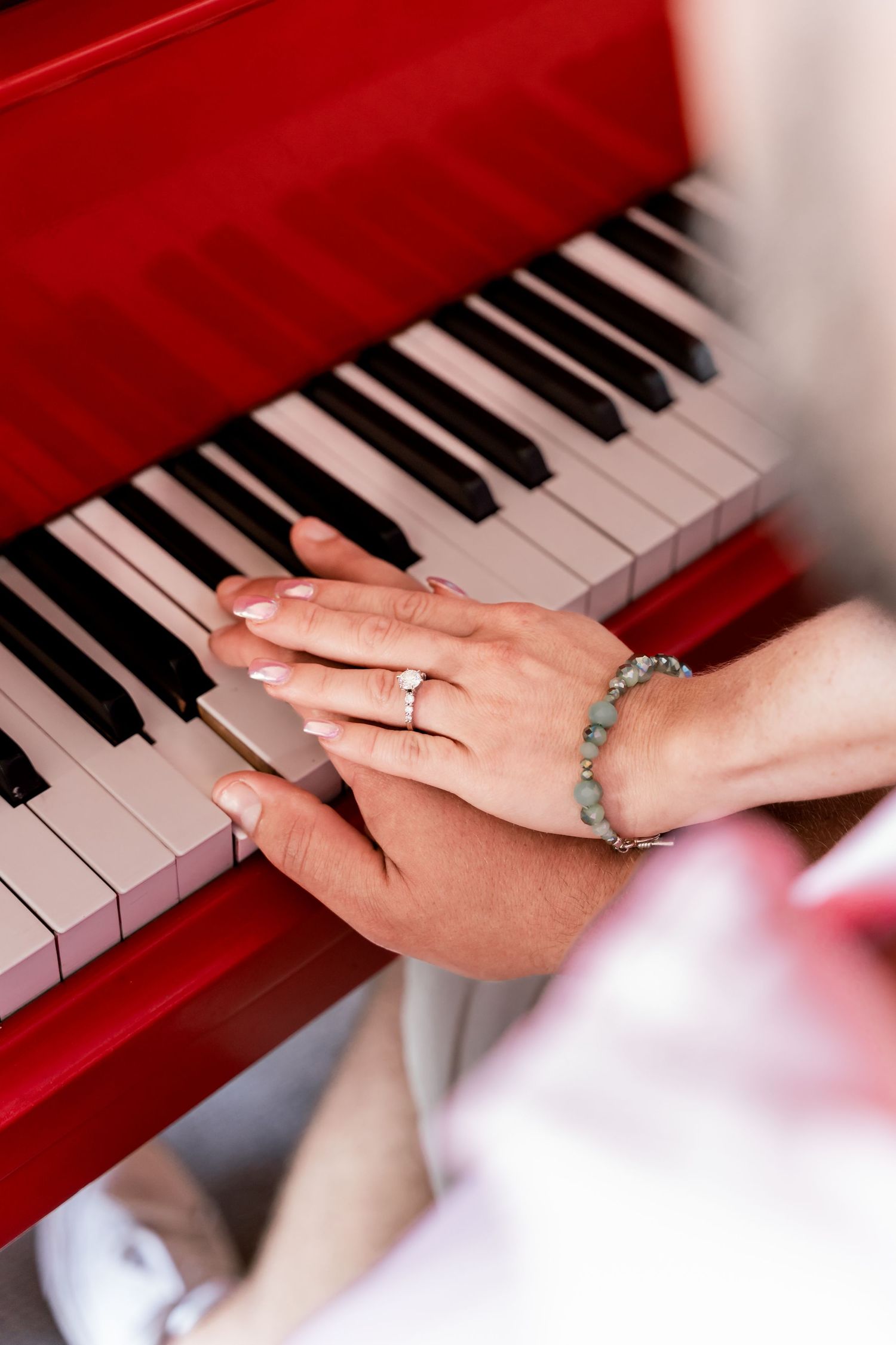 Hands wearing a diamond ring and jade bracelet rest gently on the keys of a bright red piano.