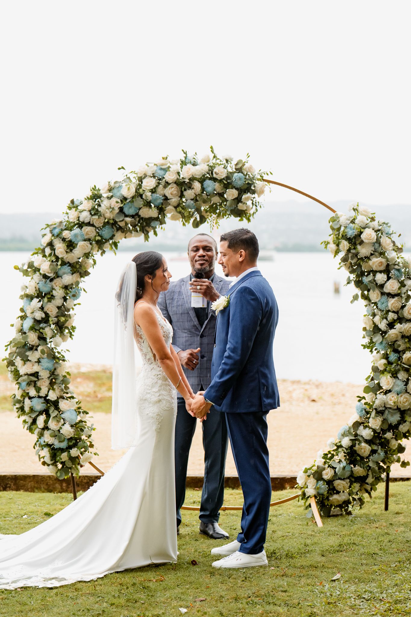 A wedding ceremony takes place under a curved floral arch with white roses overlooking a serene lake.