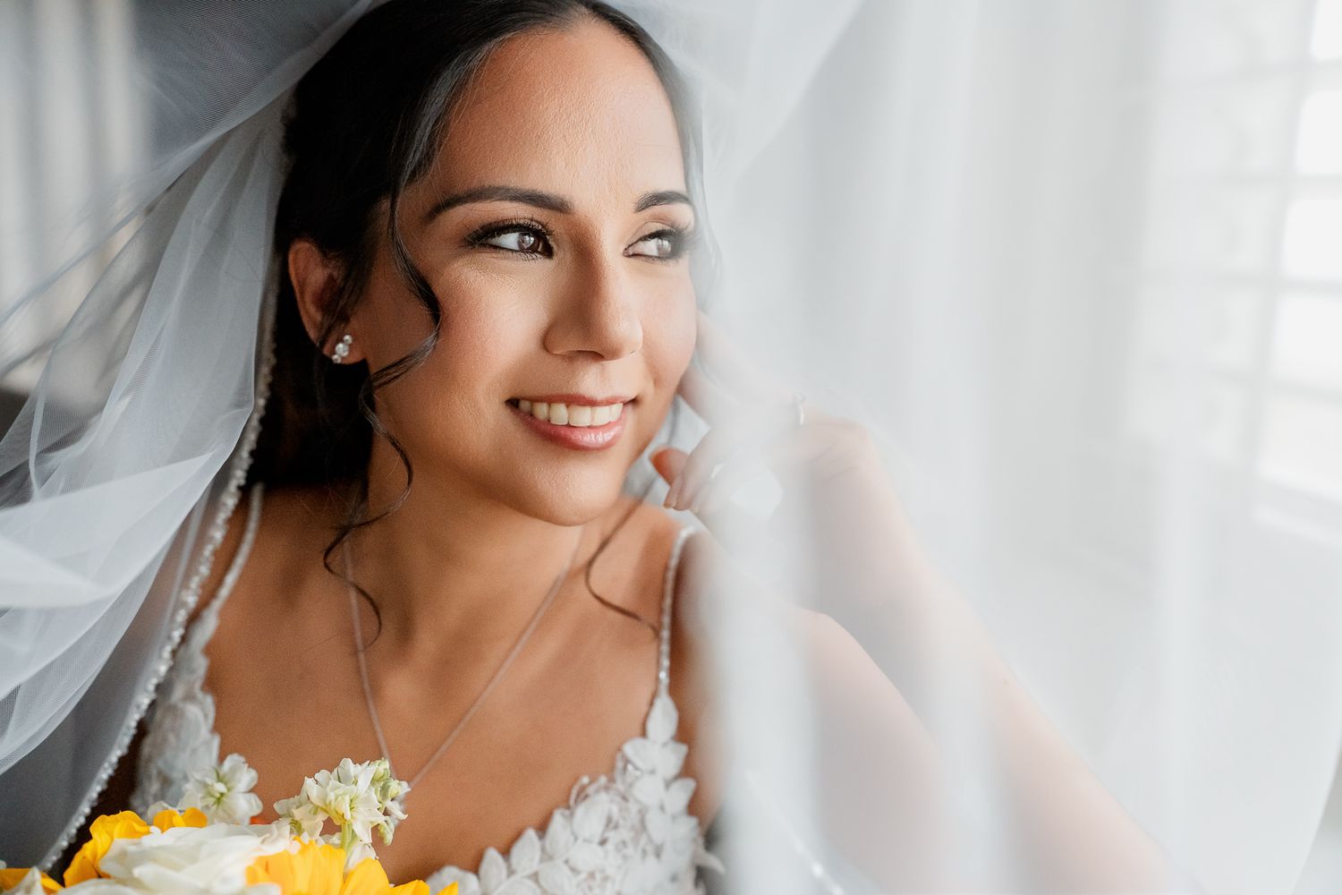 Radiant bride wearing a white lace wedding dress holds yellow flowers against a light curtained background.