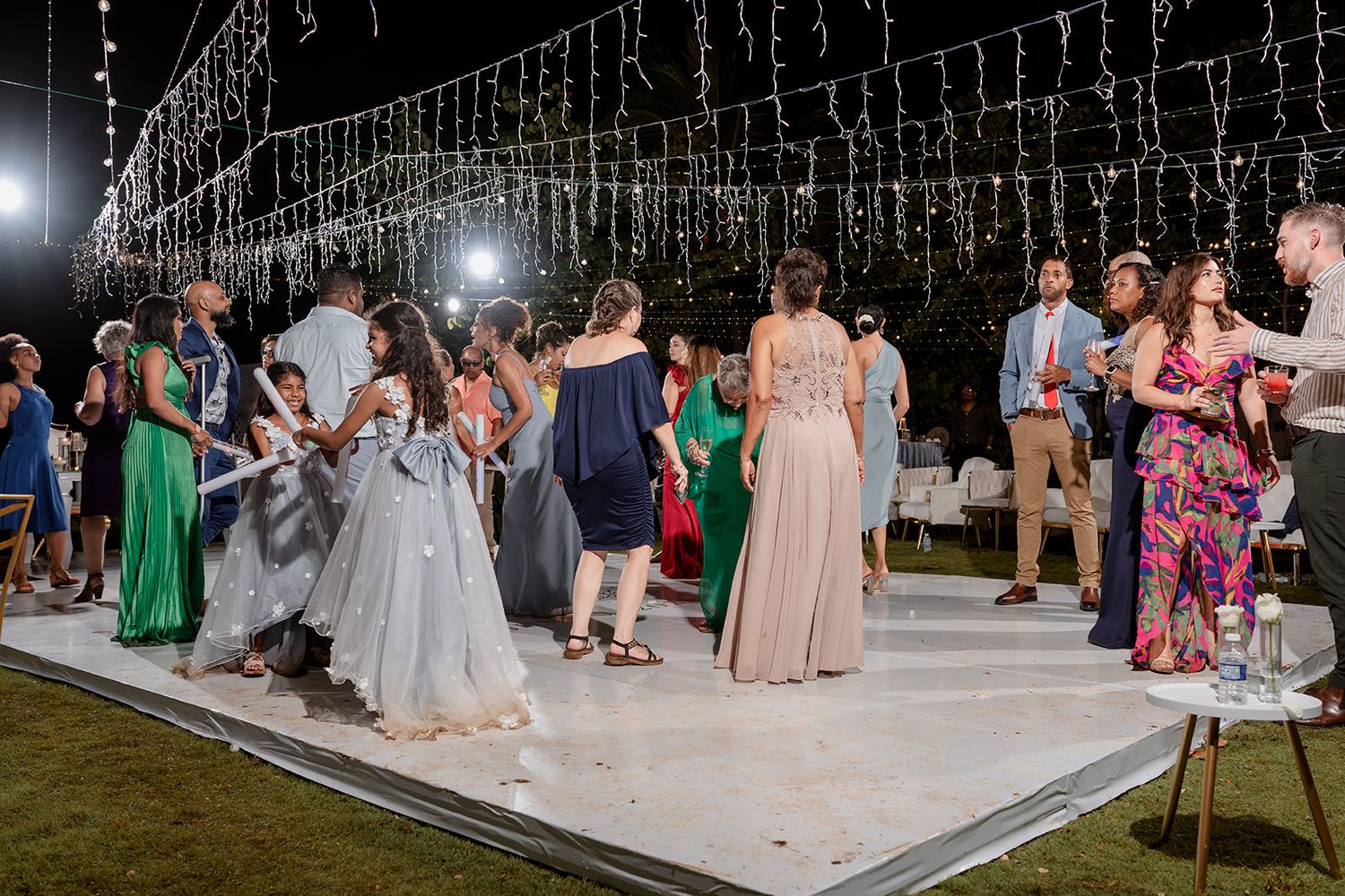 Guests dance at an outdoor evening wedding reception under string lights on a white dance floor.
