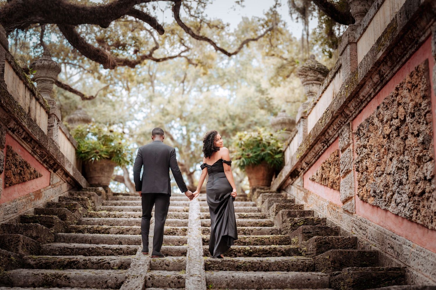 A couple walks up ornate stone steps with decorative red railings under overhanging trees at sunset.