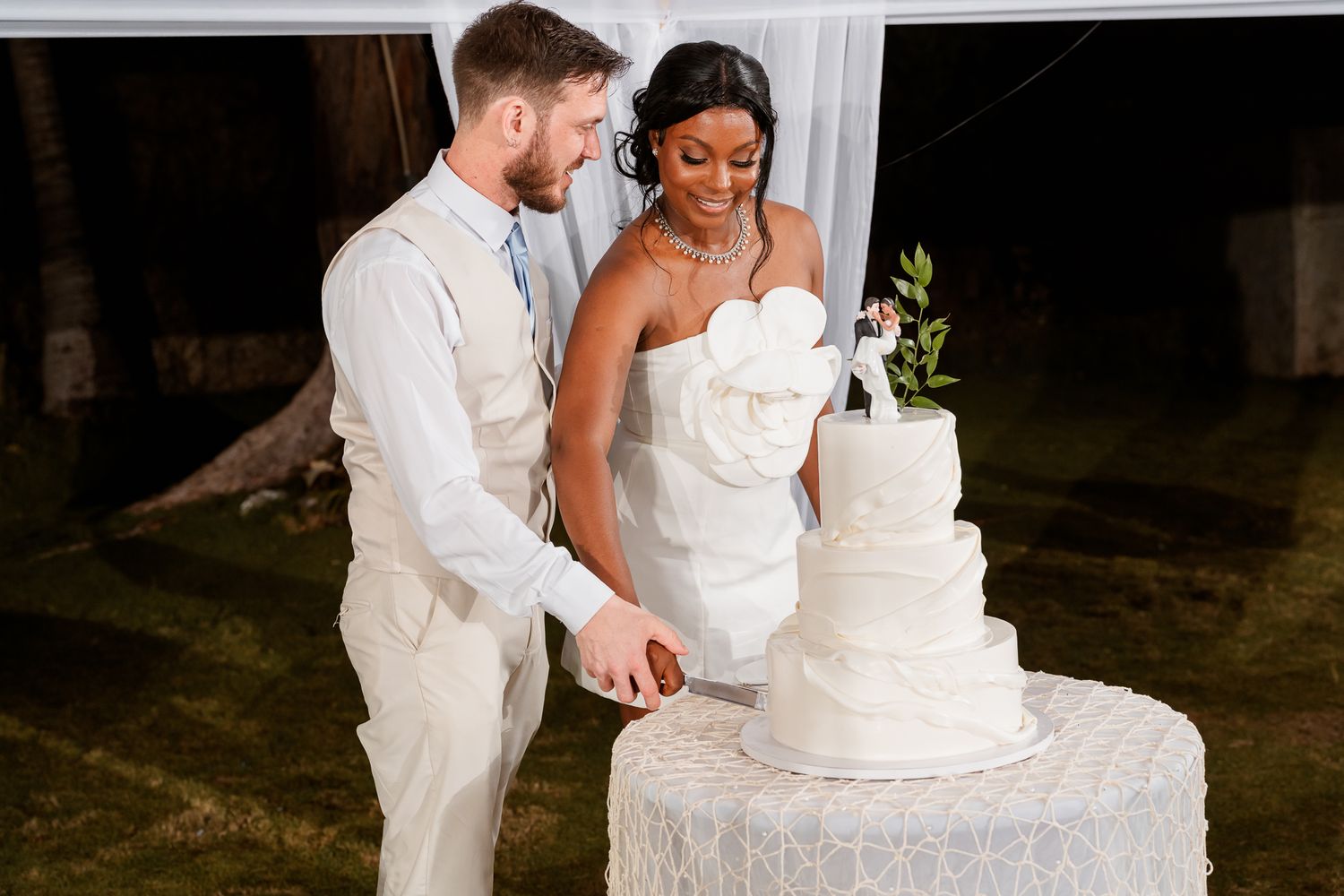 Interracial couple cutting white tiered wedding cake, bride in white dress, groom in tan suit, outdoor reception.