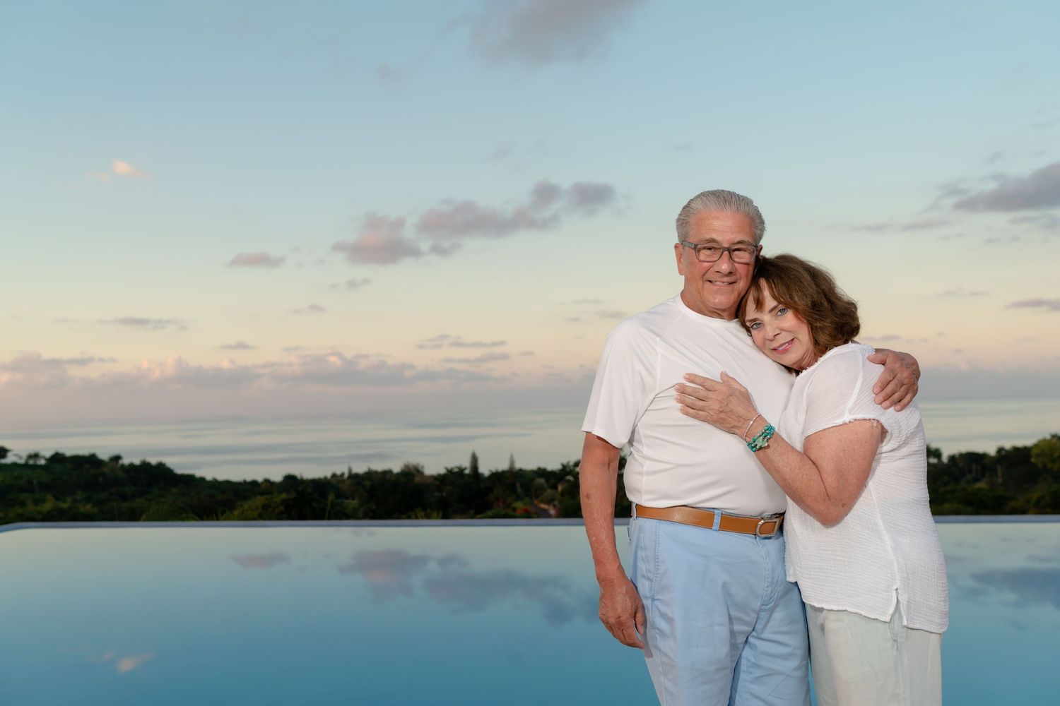 Loving elderly couple embraces while standing near tranquil waterfront at sunset with pastel sky in background.