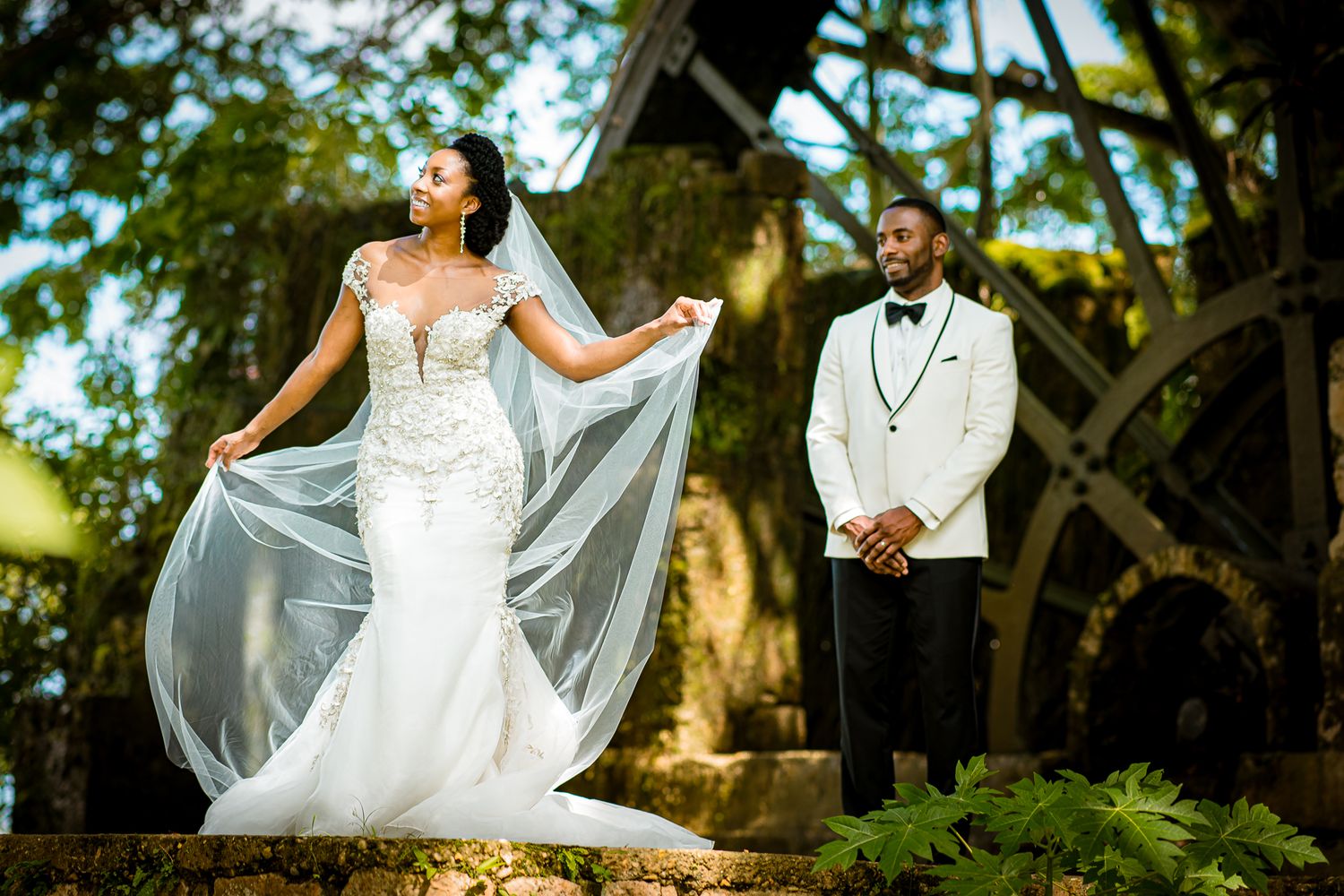 Bride in lace mermaid gown with flowing veil stands beside groom in white tuxedo at outdoor wedding ceremony Tryall Gold Club.