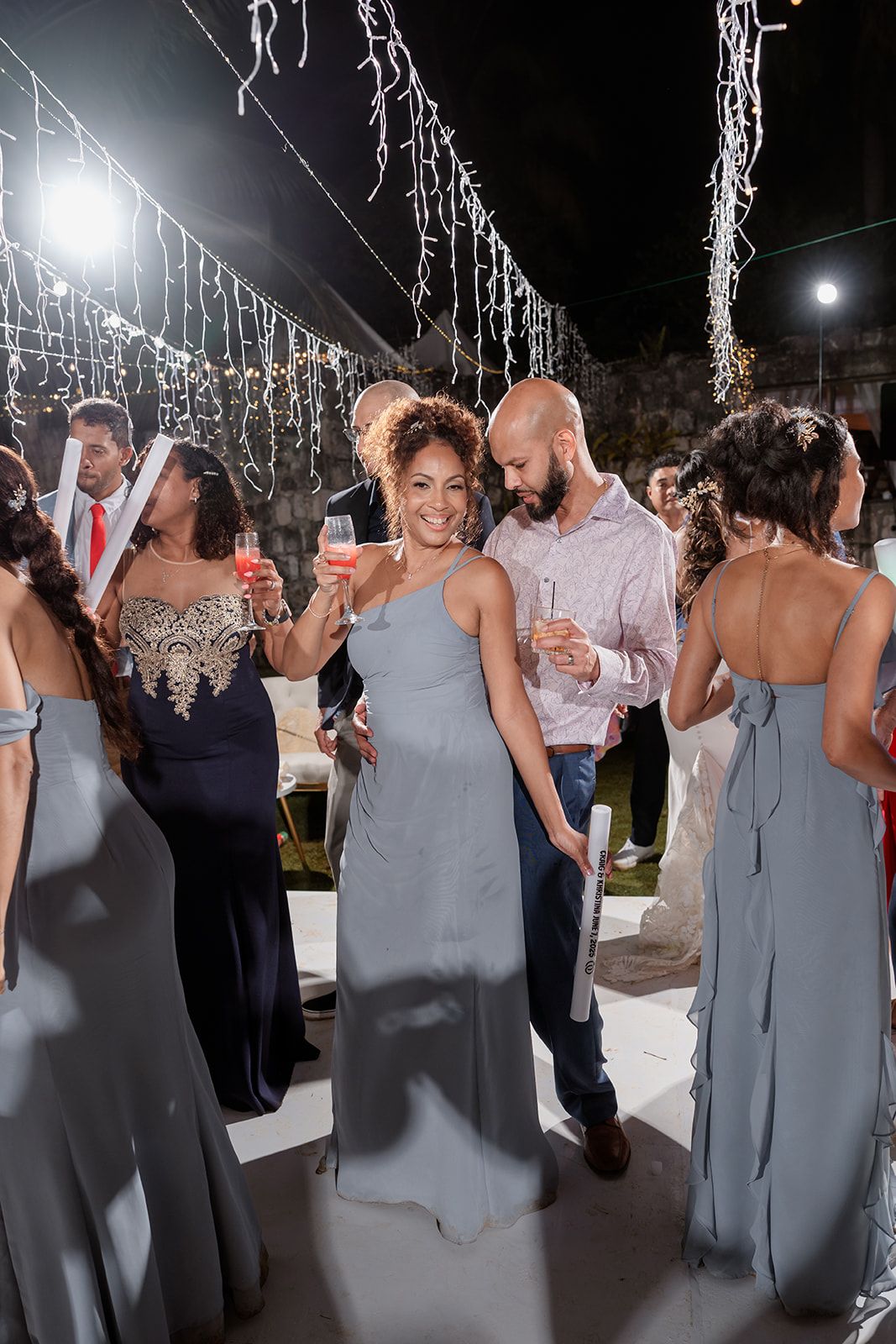 Wedding guests in matching slate-blue dresses dance and celebrate on a festively lit outdoor dance floor at night.