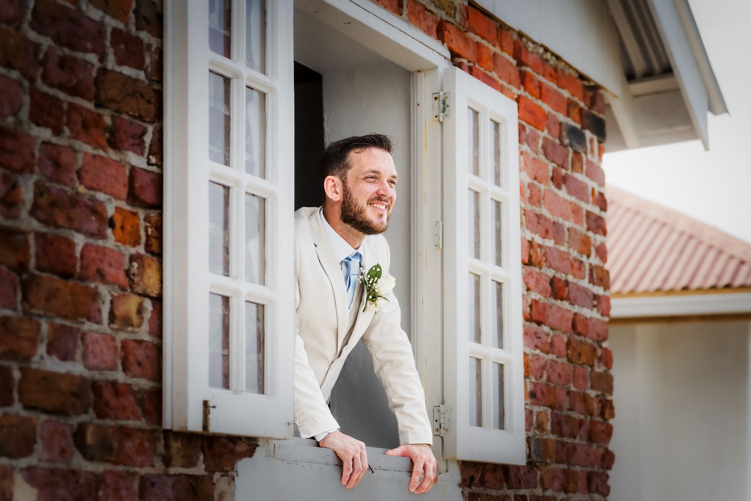 Smiling groom in beige suit with boutonniere leans out of white French doors on brick building.