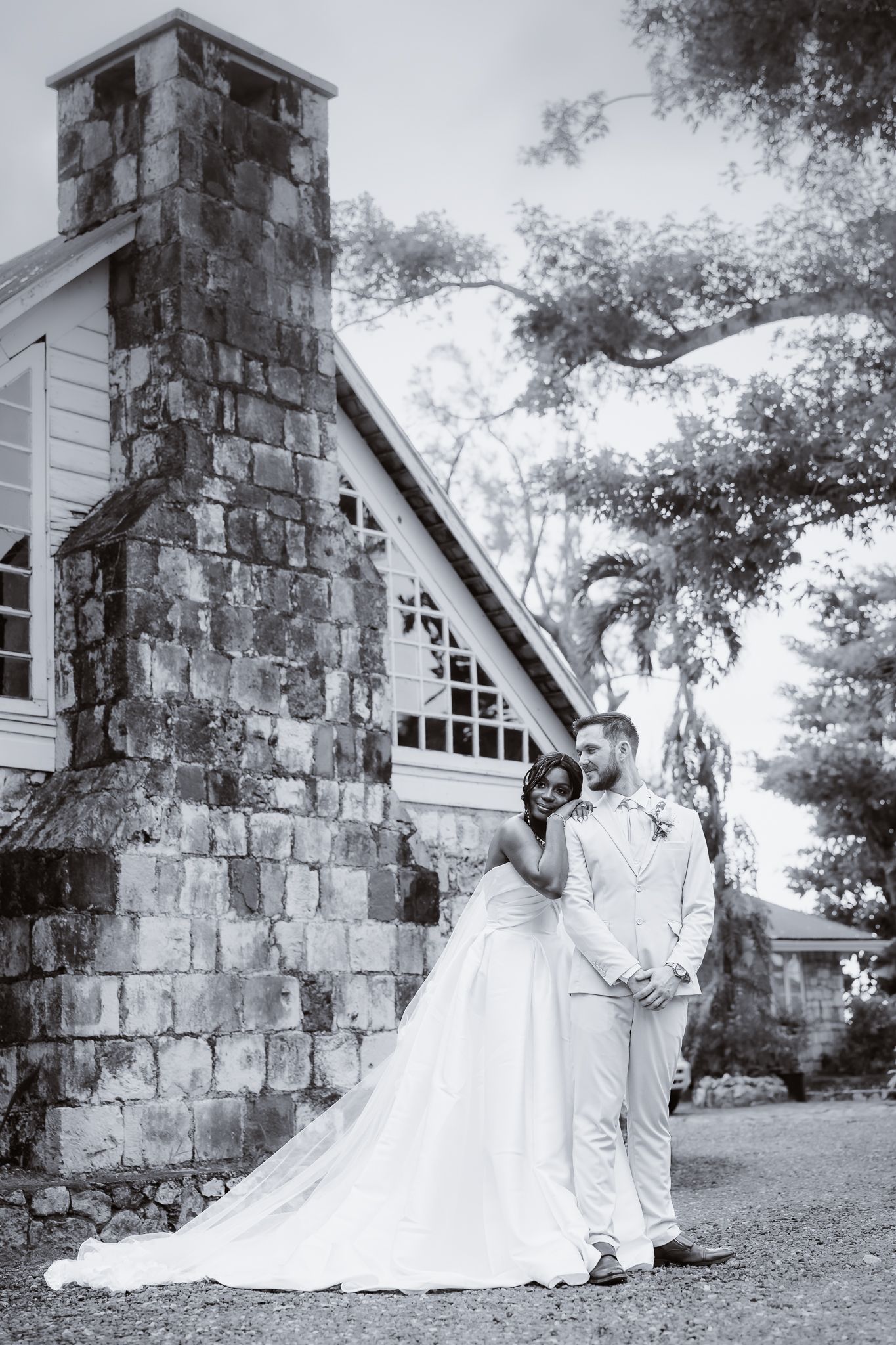 Black and white photo of bride and groom posing romantically by a stone chapel surrounded by lush trees.